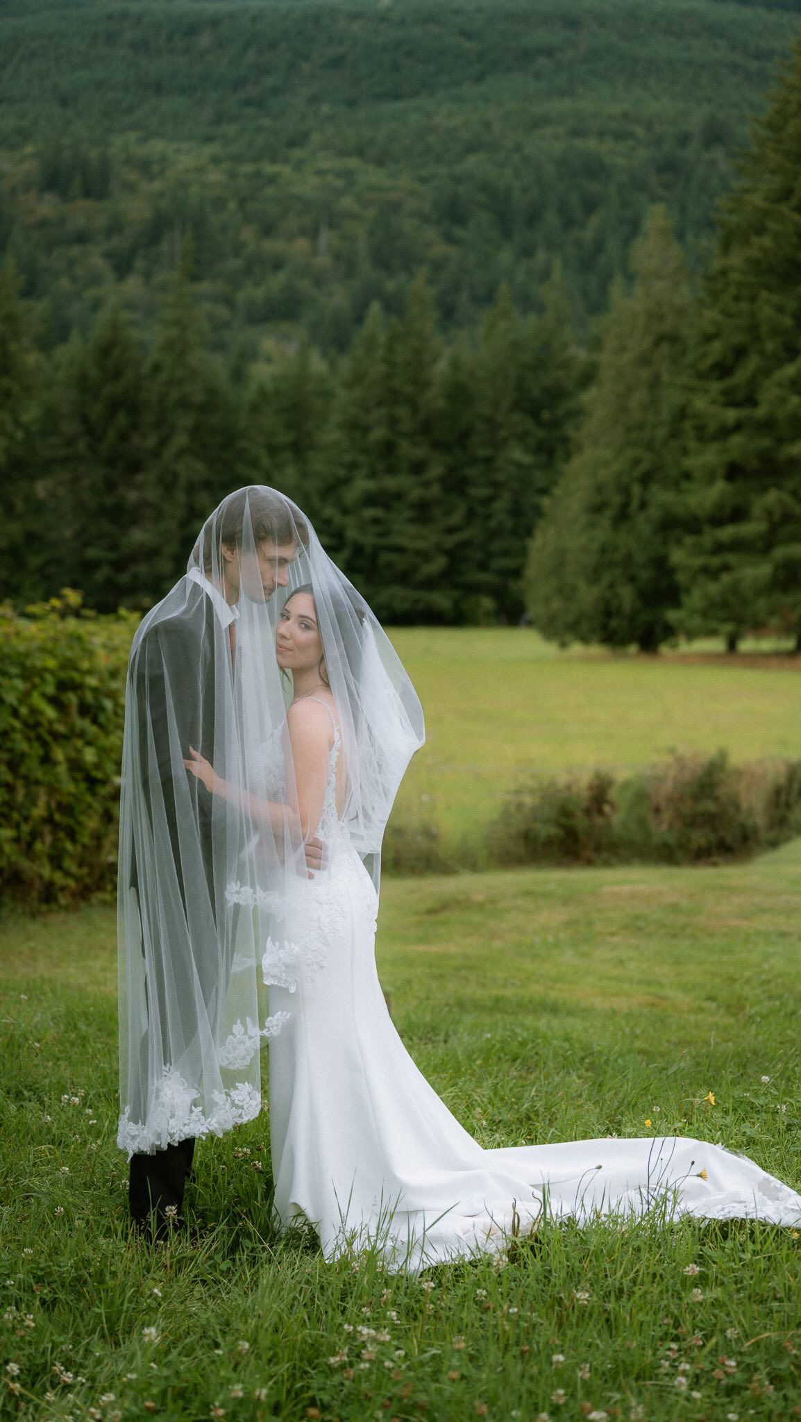 Bride and groom wrapped together in a long veil on a green lawn