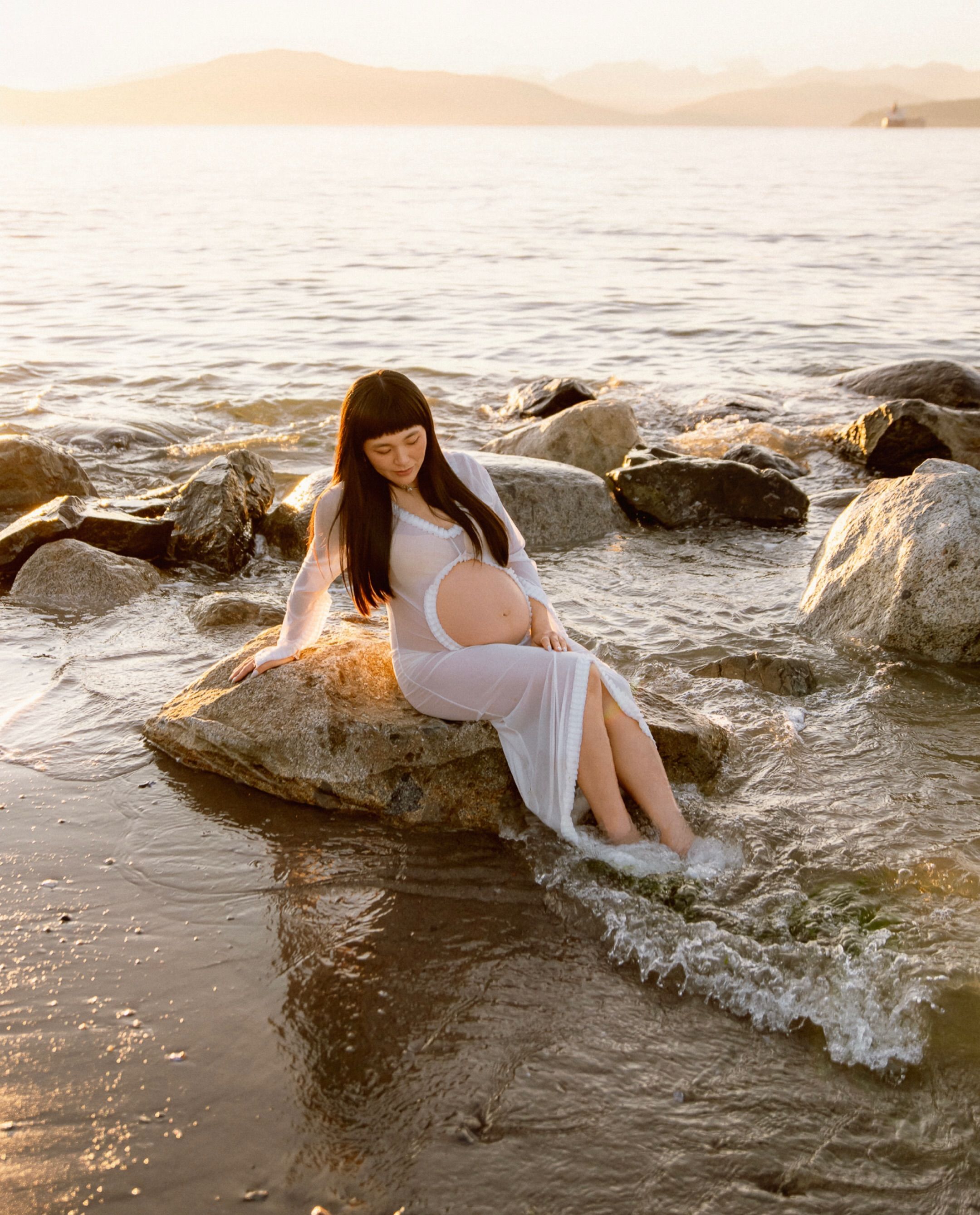 Expectant mother in a white dress seated on shoreline rocks at sunset