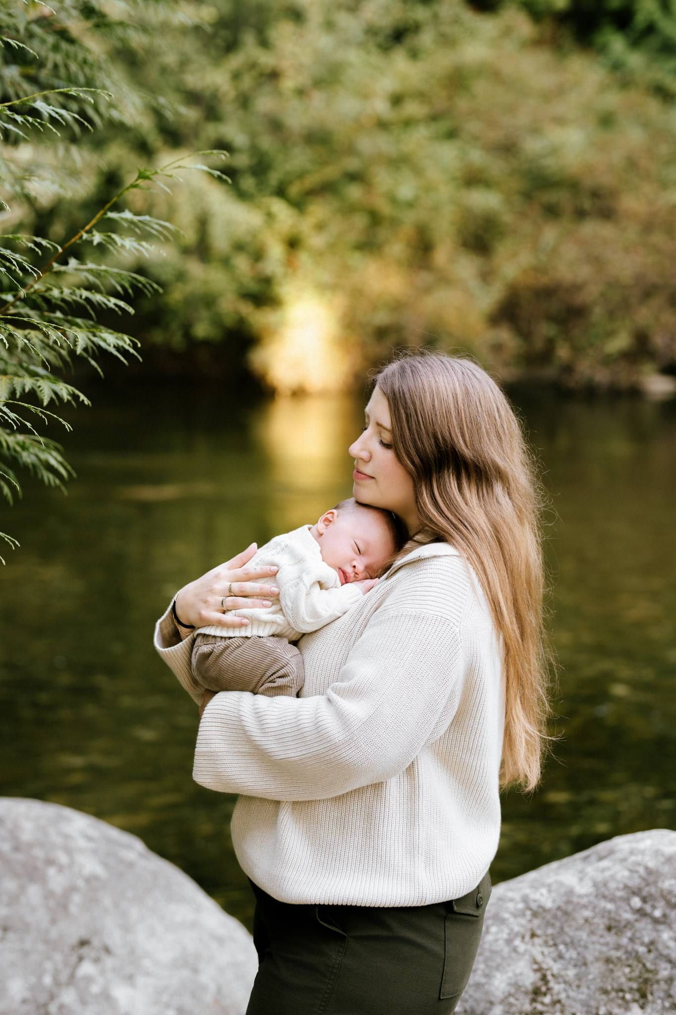 Mother in a white sweater holding her newborn beside a forest river