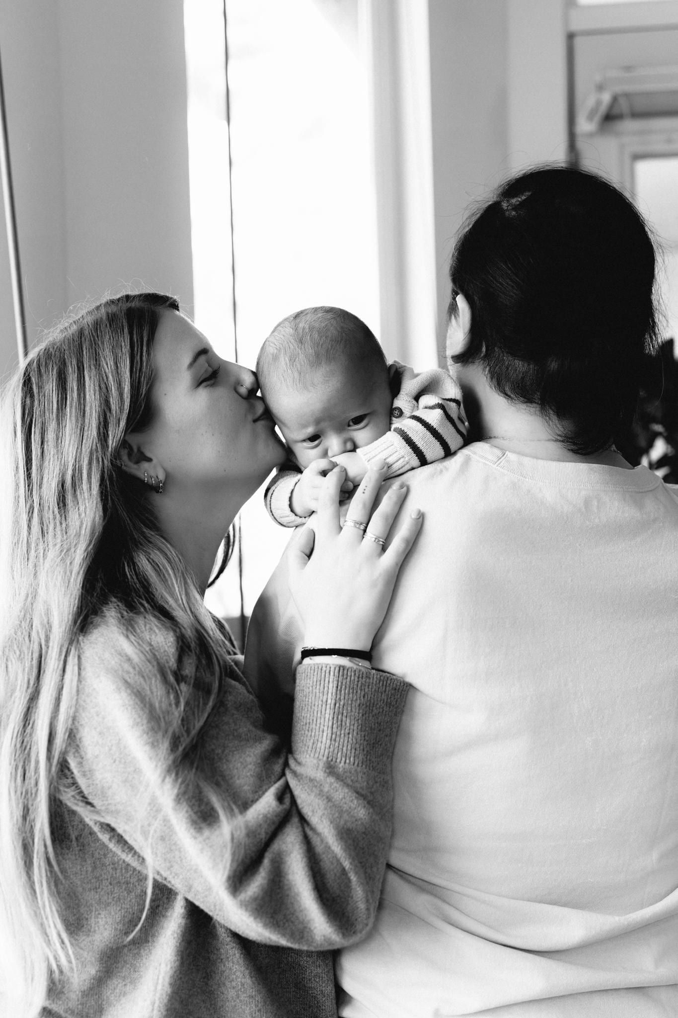 Black-and-white photo of a mother kissing her newborn over the father's shoulder