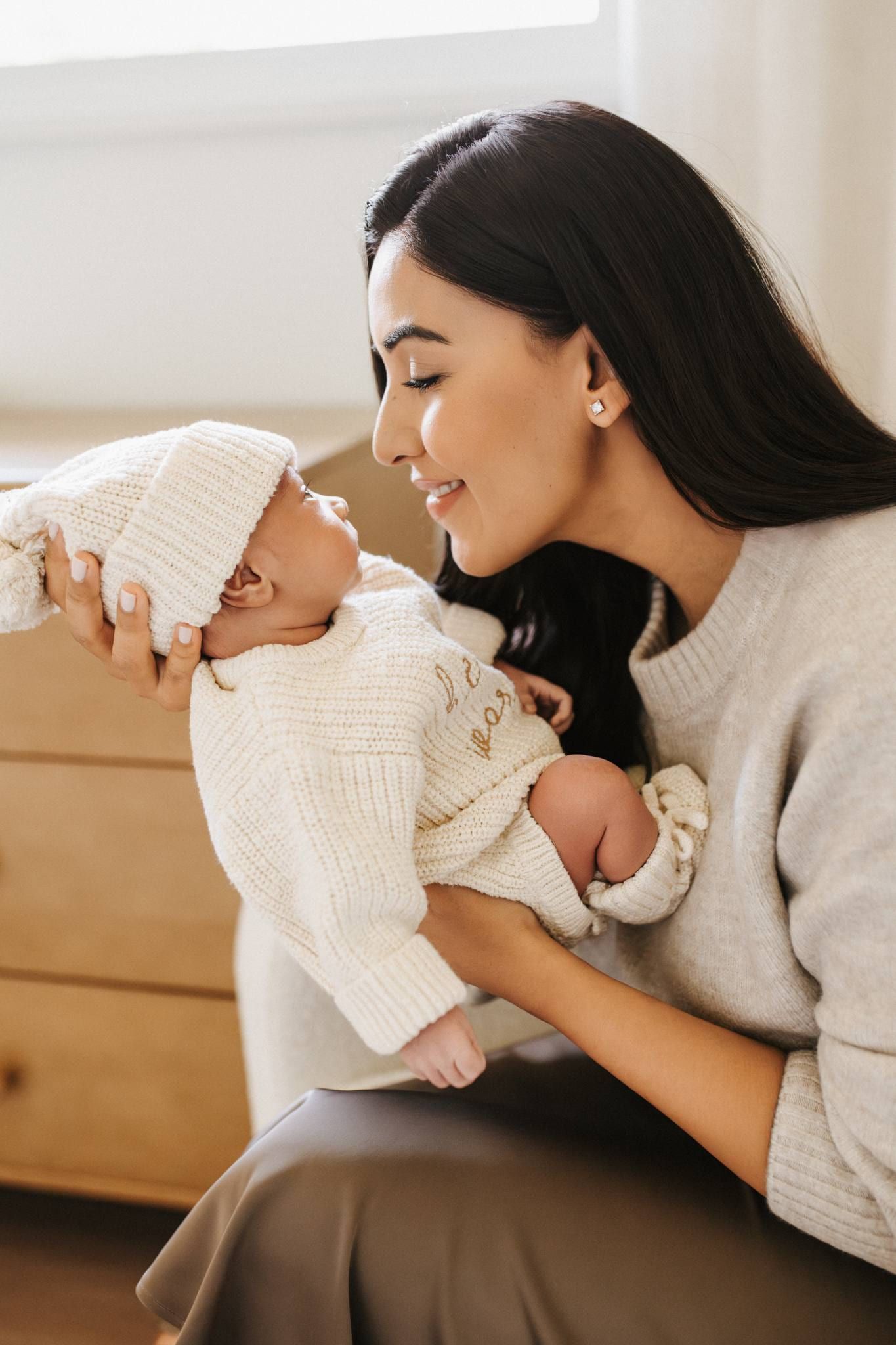Mother in a grey sweater smiling down at her newborn in a knitted outfit