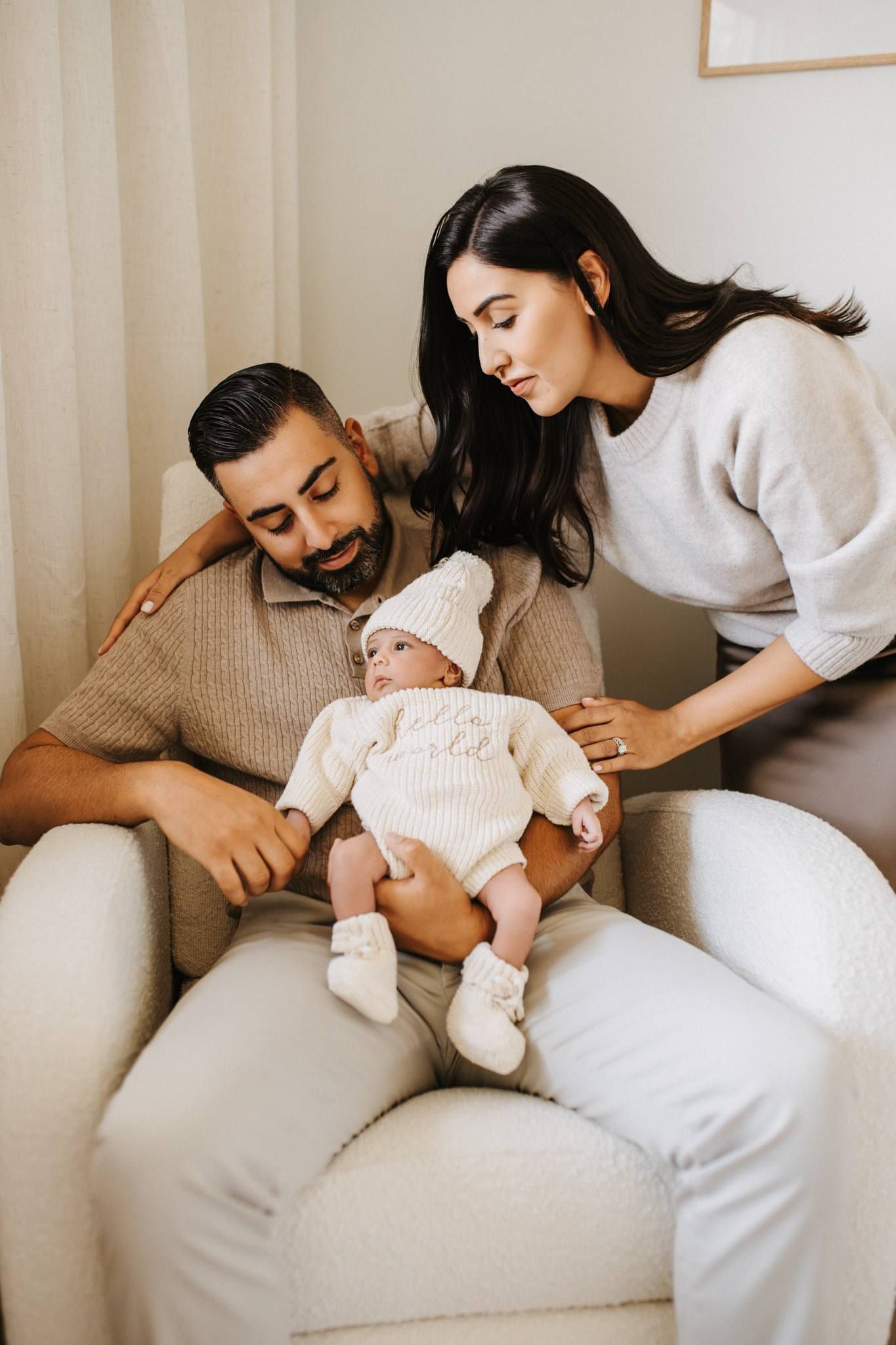 Parents on an armchair gazing at their newborn in a white knitted hat