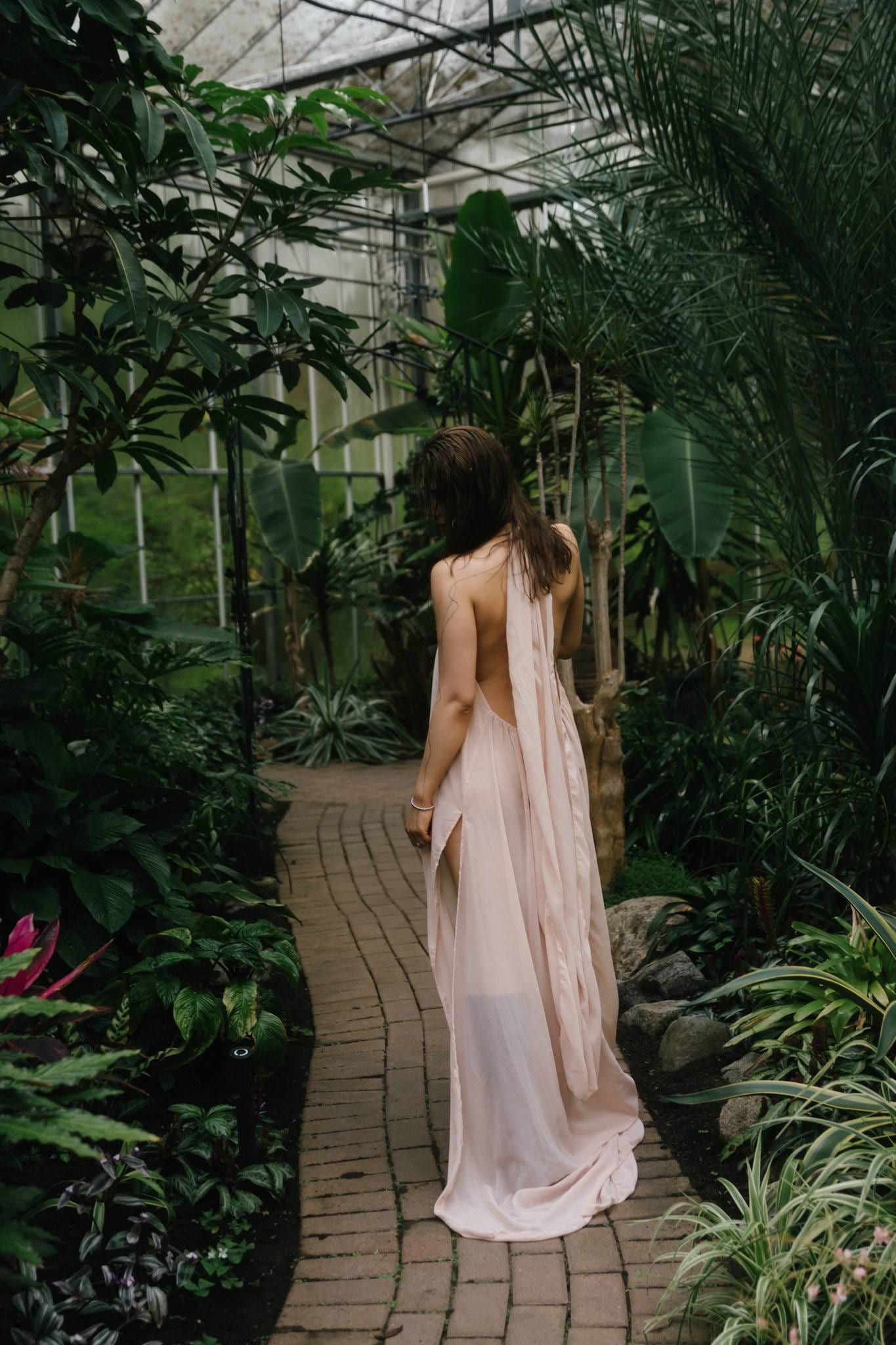 Woman in a blush backless gown on a brick path inside a lush conservatory
