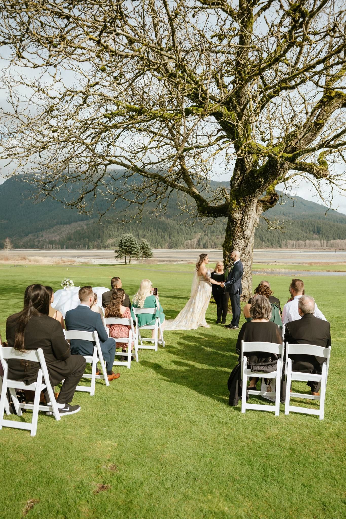 Guests seated on white chairs before a bare oak tree at a riverside ceremony