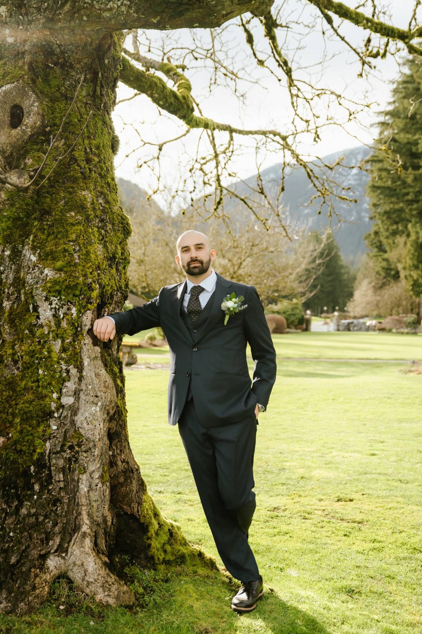 Groom in a navy suit leaning against a mossy tree