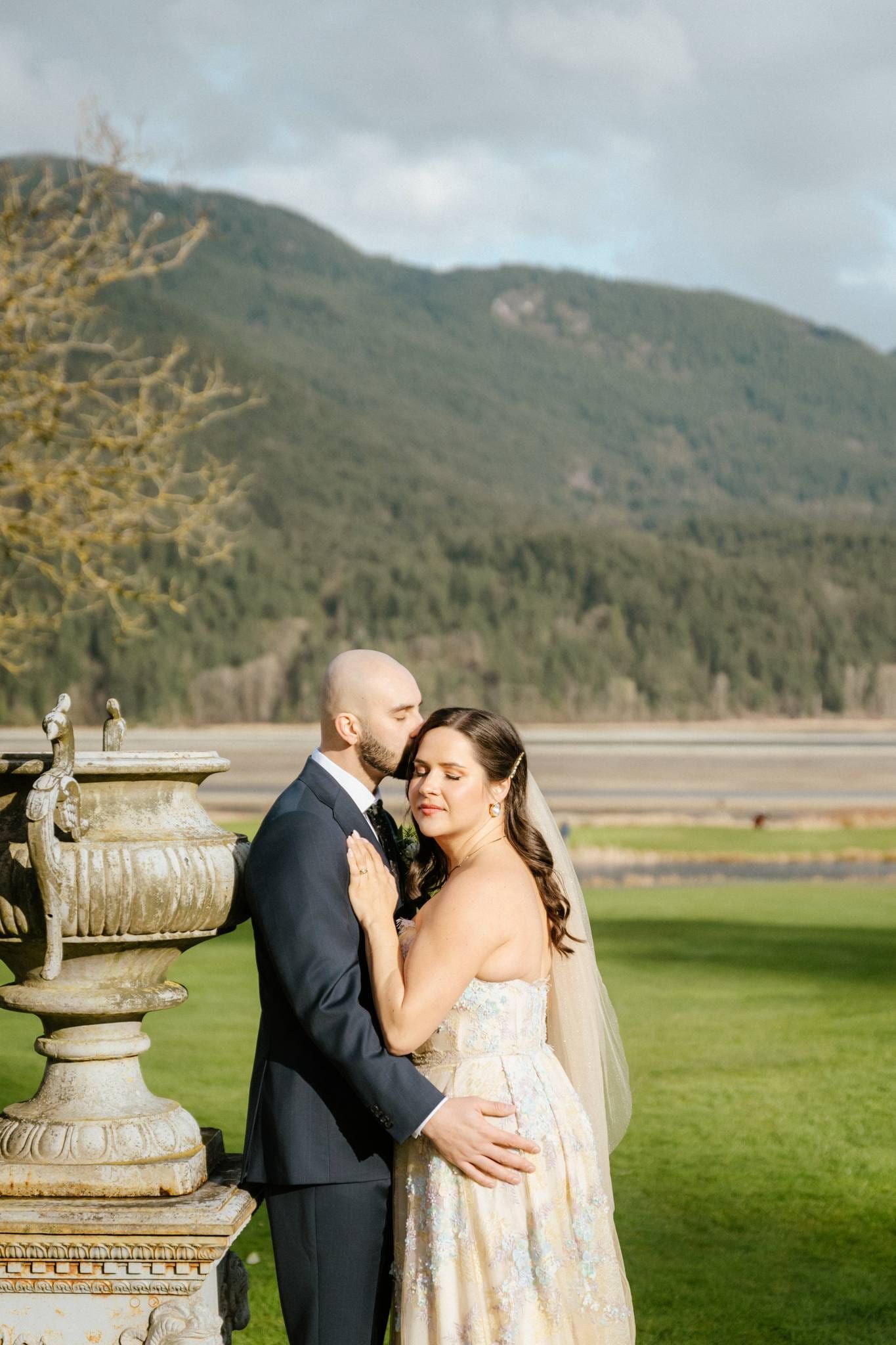 Groom holding the bride close beside a stone urn, mountains behind