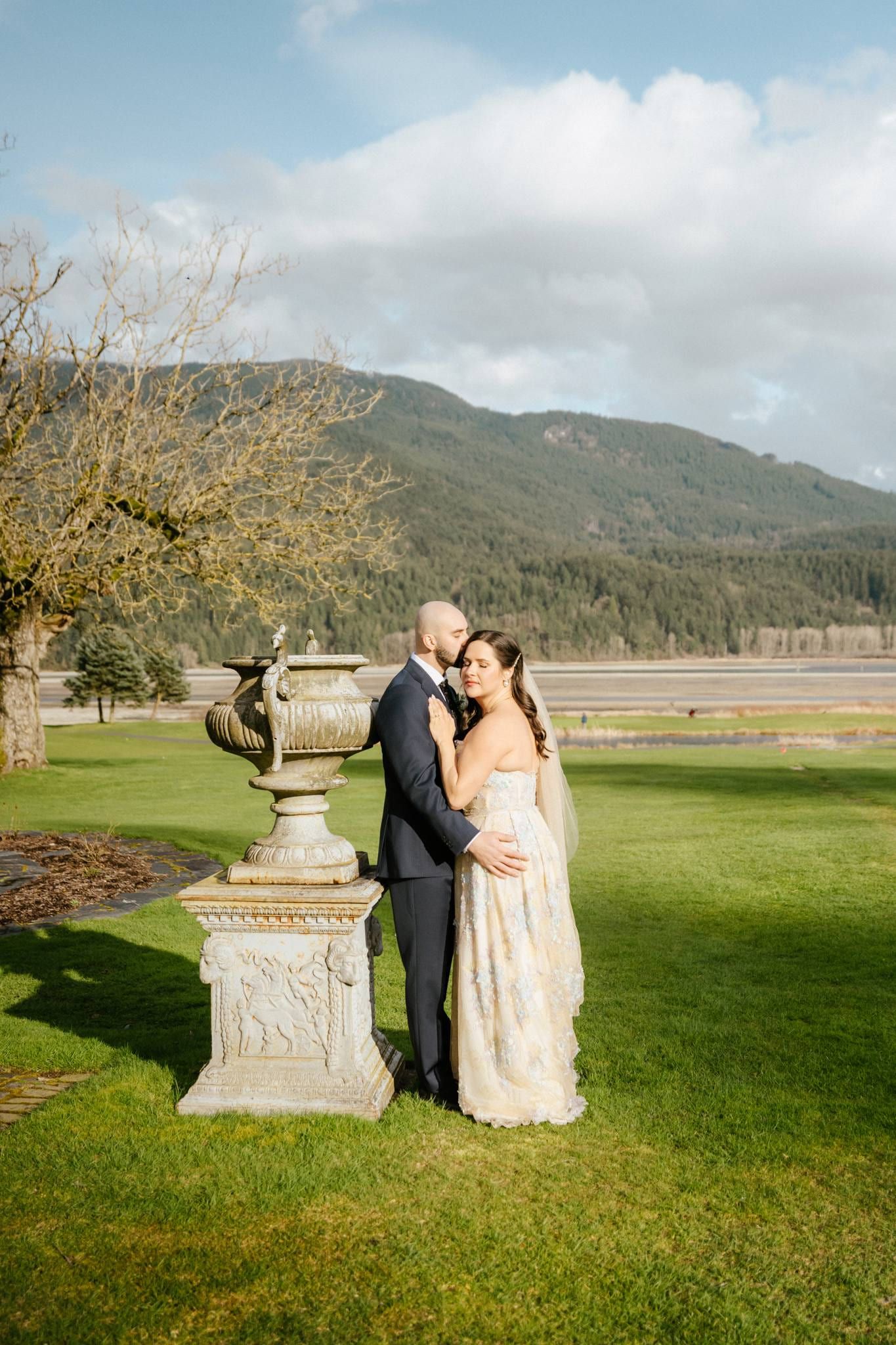 Couple posing beside a stone urn on the lawn with mountains behind
