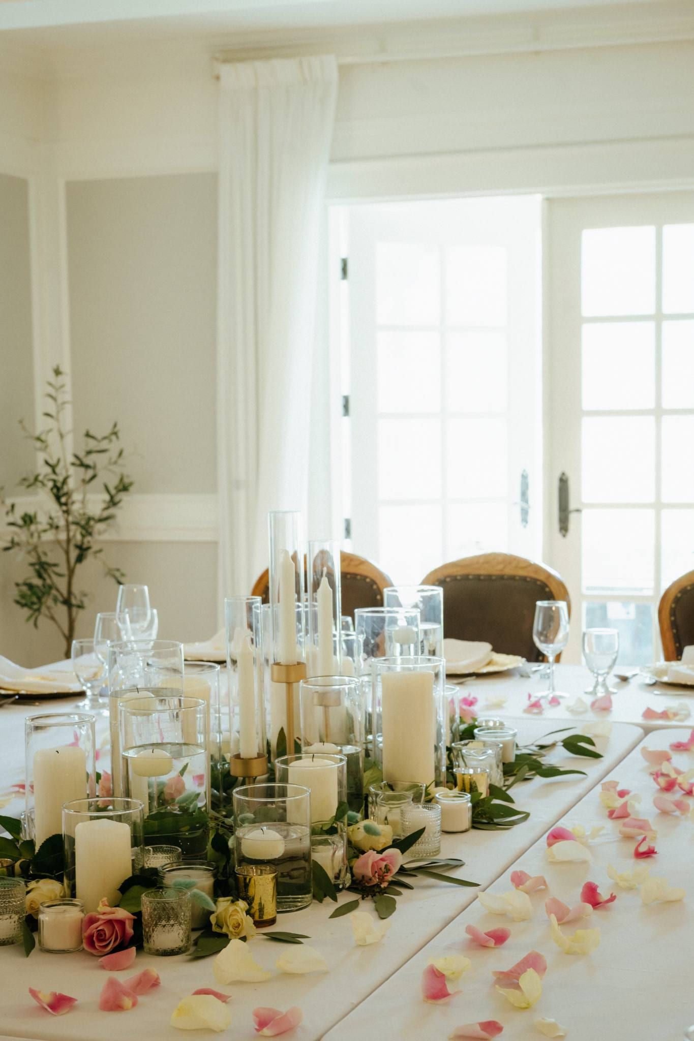 Reception table set with candles and rose petals by tall windows