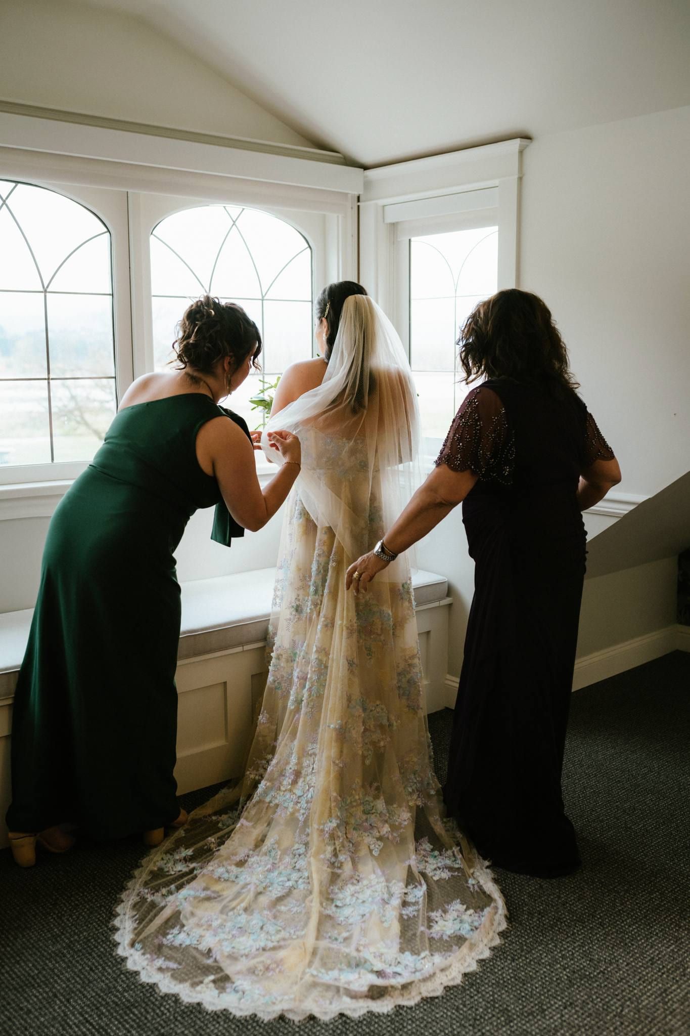 Bride's mother and friend fastening her veil in a bright window seat