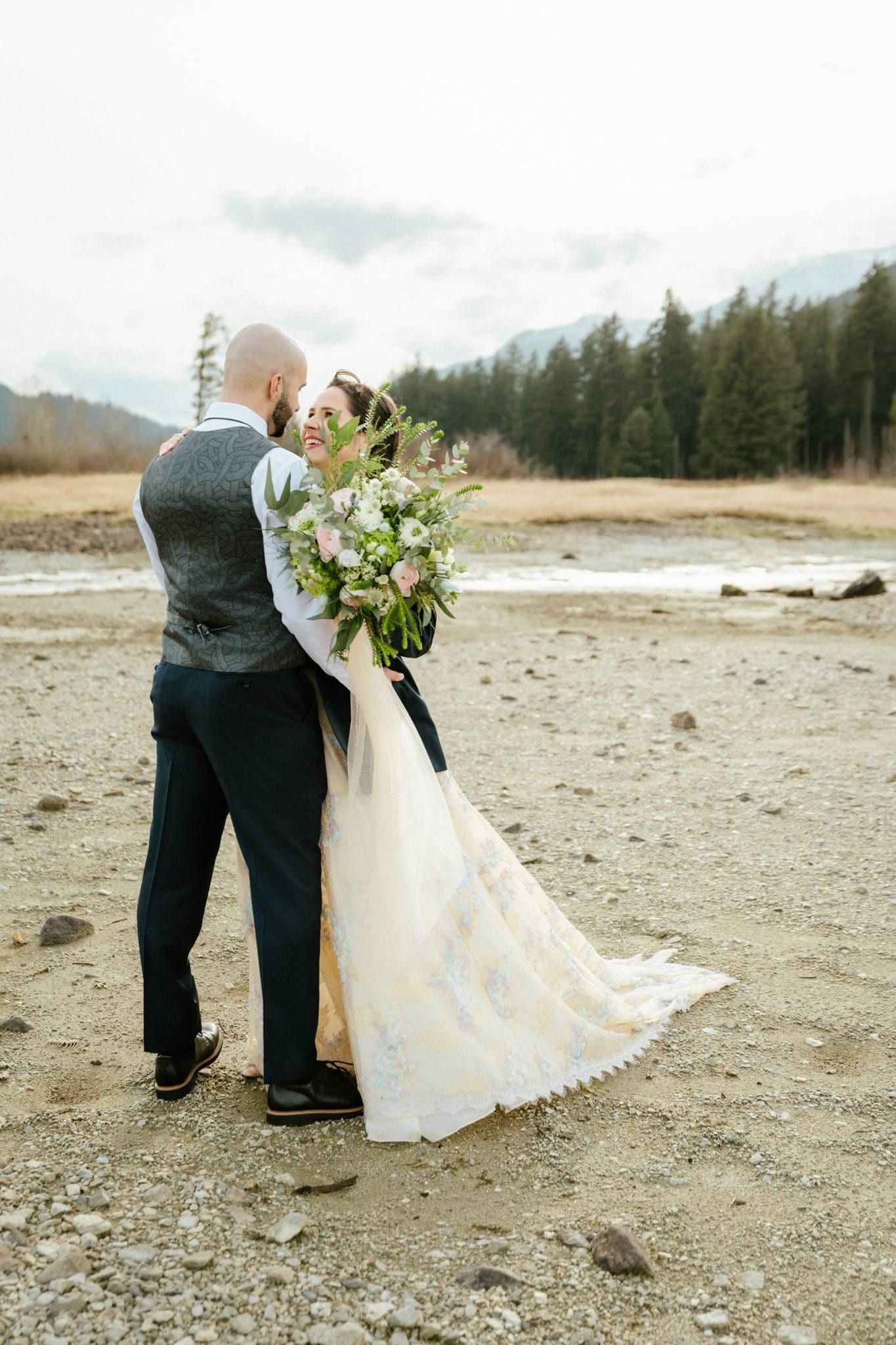 Groom holding the bride, her bouquet over his shoulder, on a gravel riverbed beneath the mountains