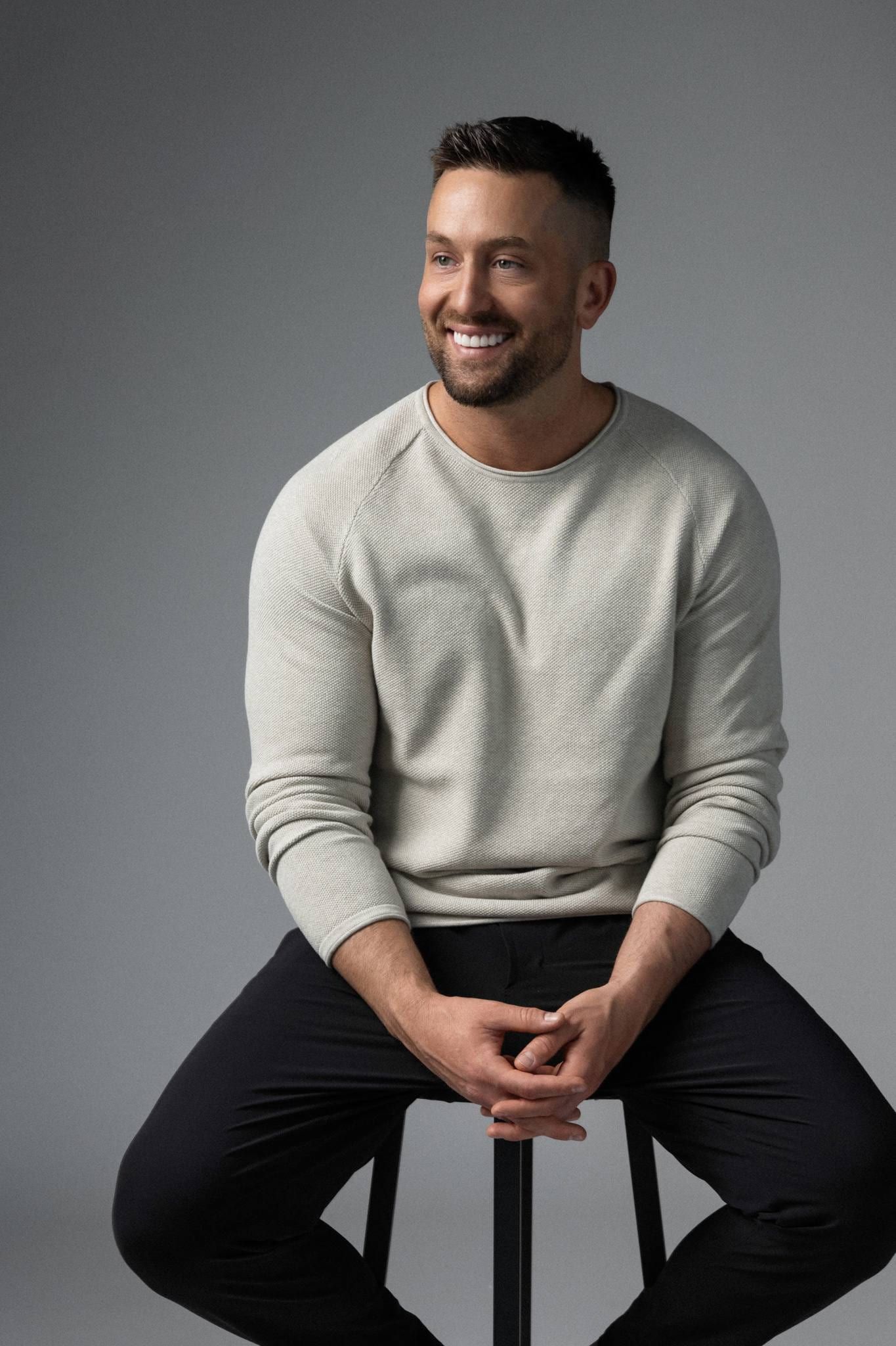 Man in a cream sweater seated on a stool, smiling
