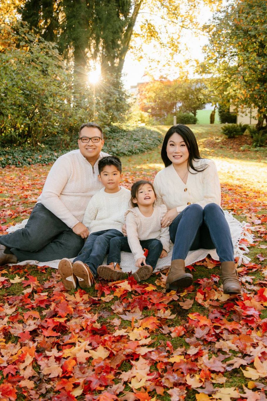Fall family photoshoot in golden light, a family of four sits on a blanket in a carpet of red autumn leaves