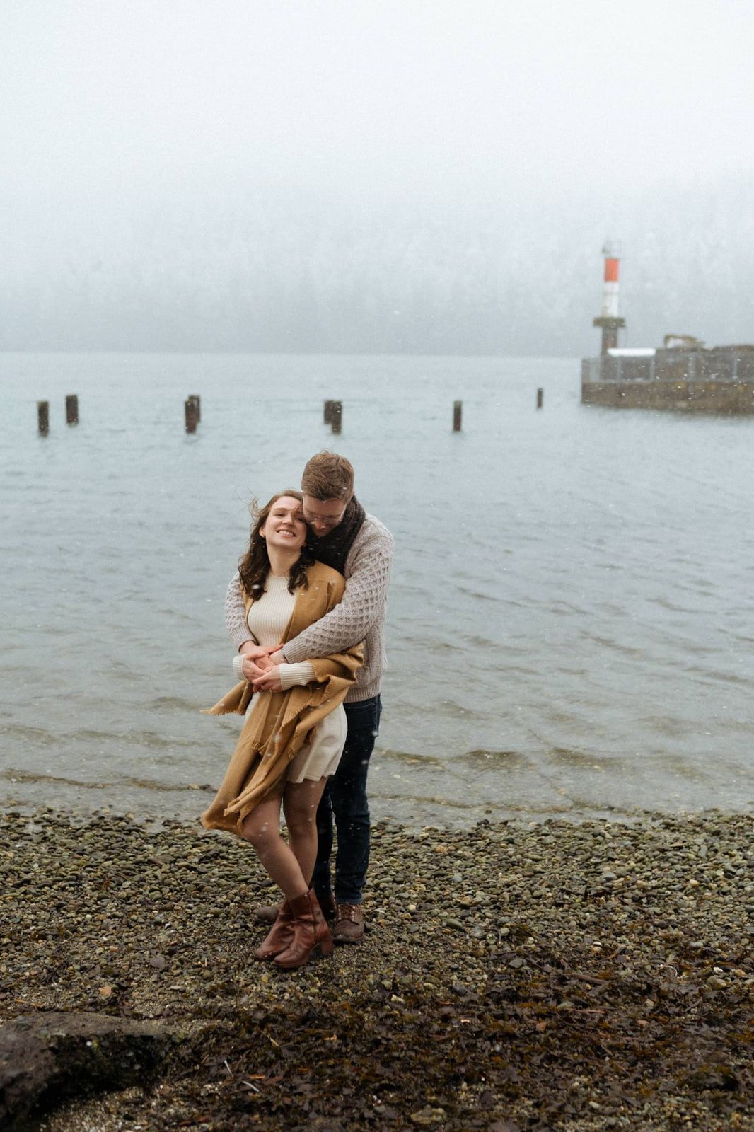 Outdoor winter engagement photos, a couple embraces on a pebble beach on a snowy day with a marker light in the distance