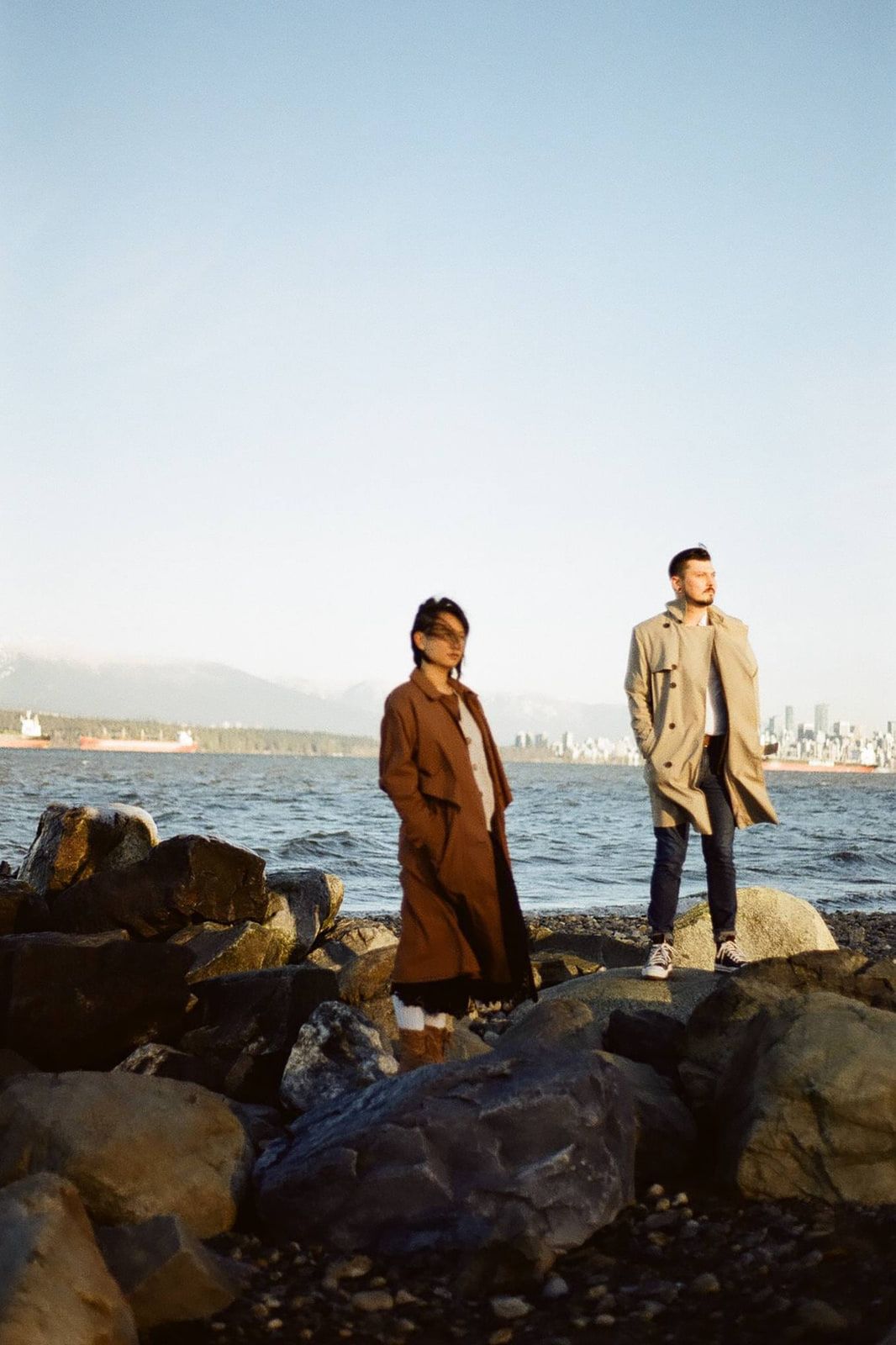 Creative engagement photos across the bay, a couple in long coats stands on waterfront rocks with a city skyline