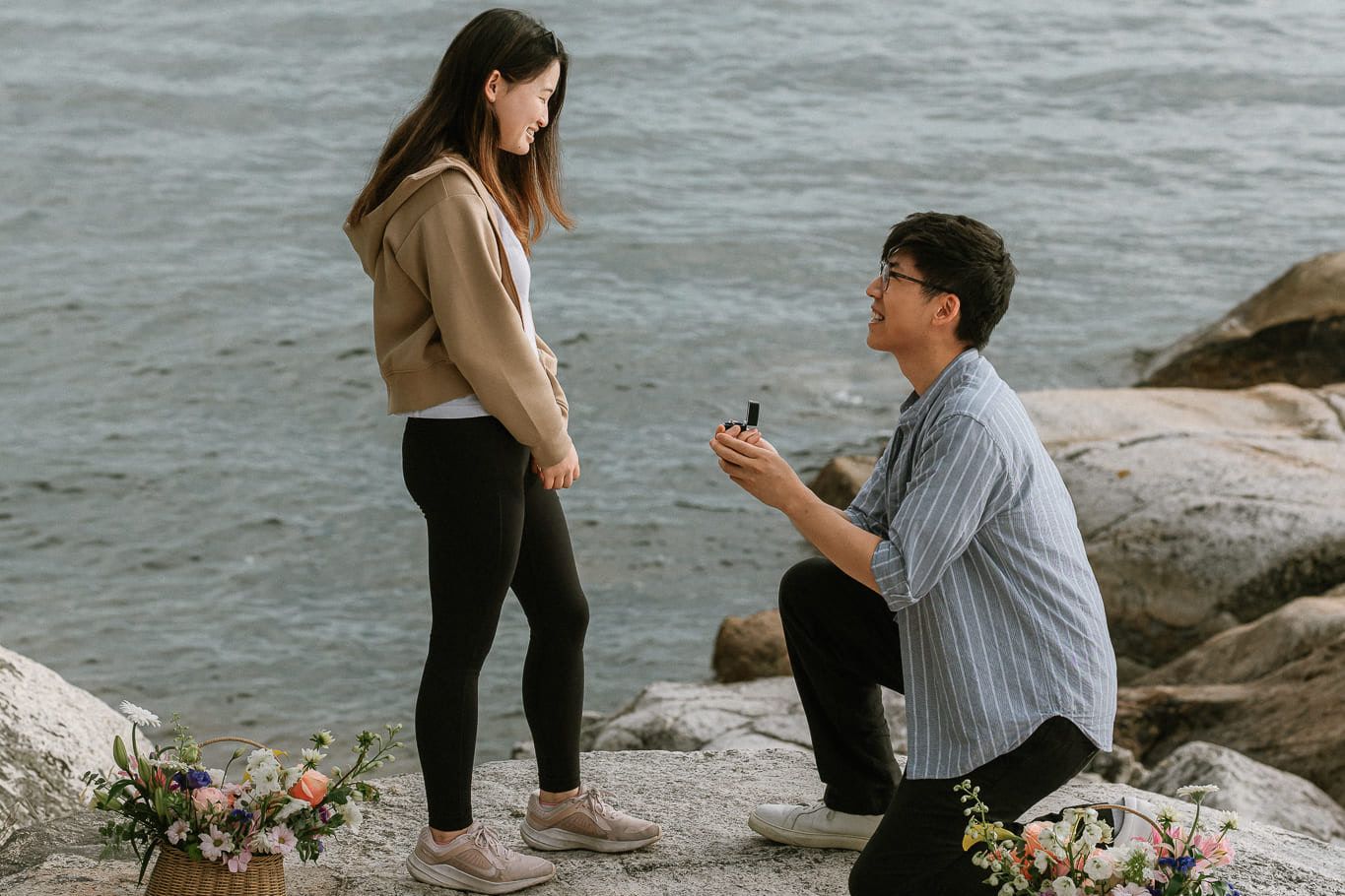 Beach engagement shoot beside two baskets of flowers, man on one knee proposing on a rocky shore