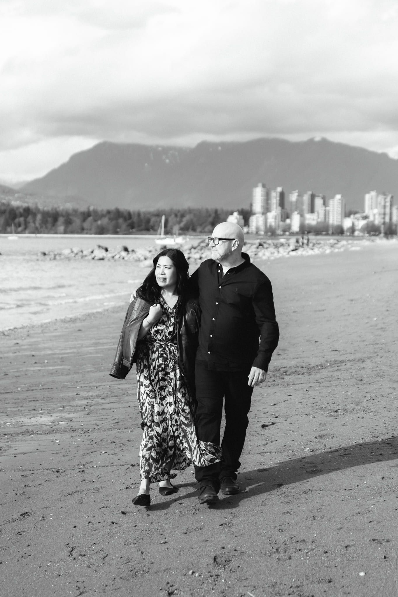 Beach engagement photos, black-and-white photo of a couple walking on a beach with the city behind