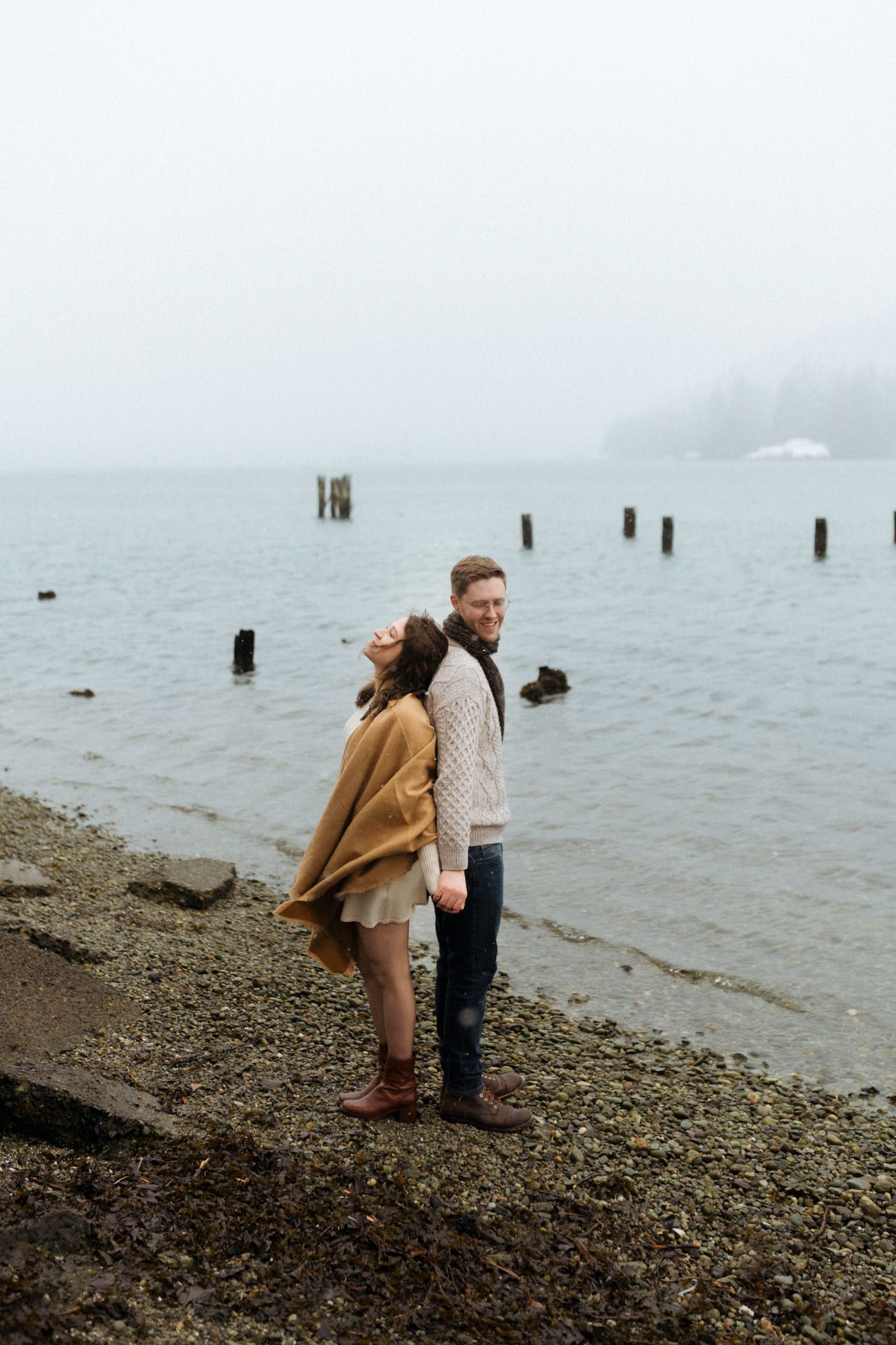 Creative engagement pictures beside old pier posts, couple embracing on a misty beach