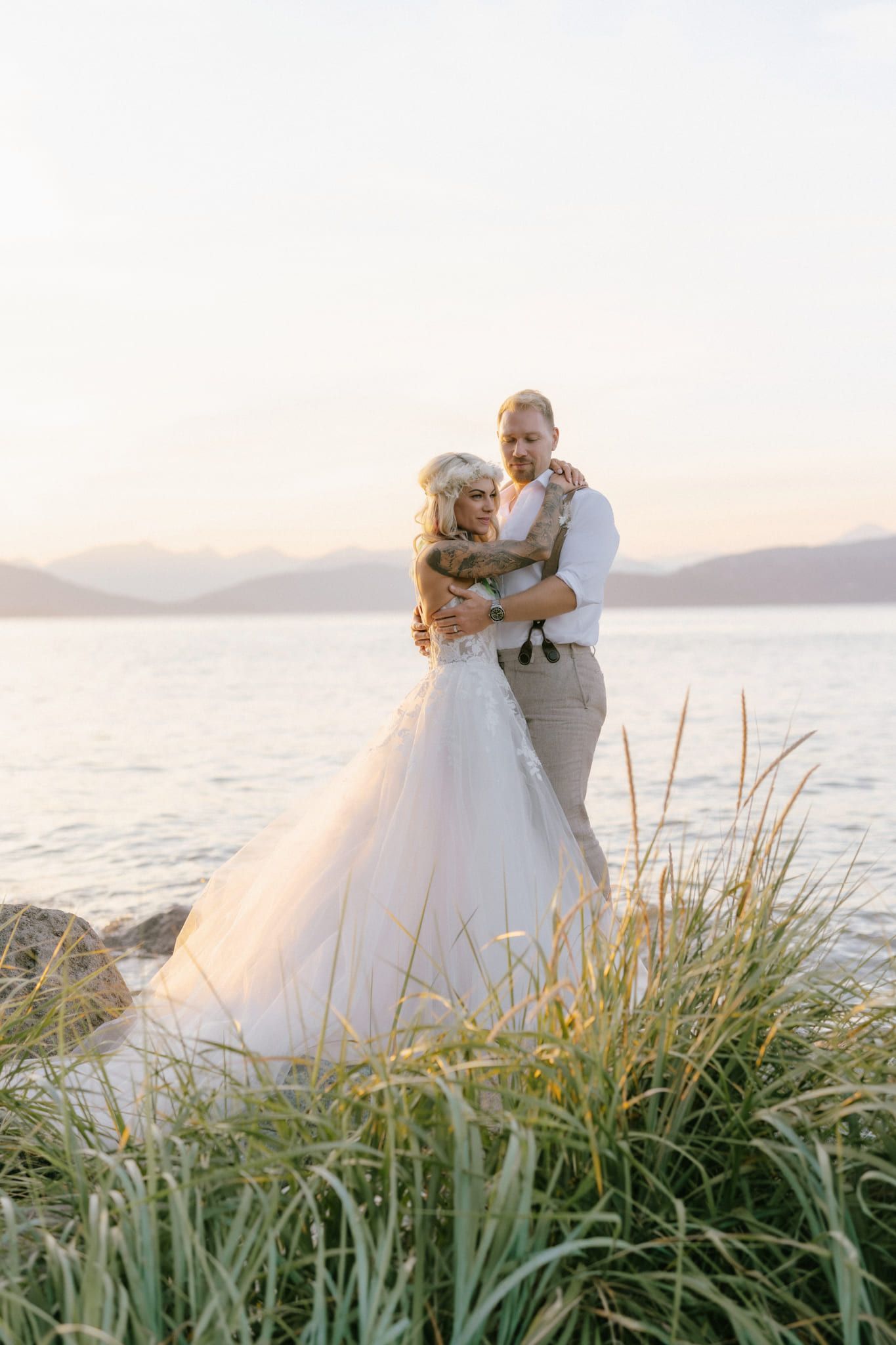 Beach wedding photography by the water at sunset, bride and groom embracing in beach grass