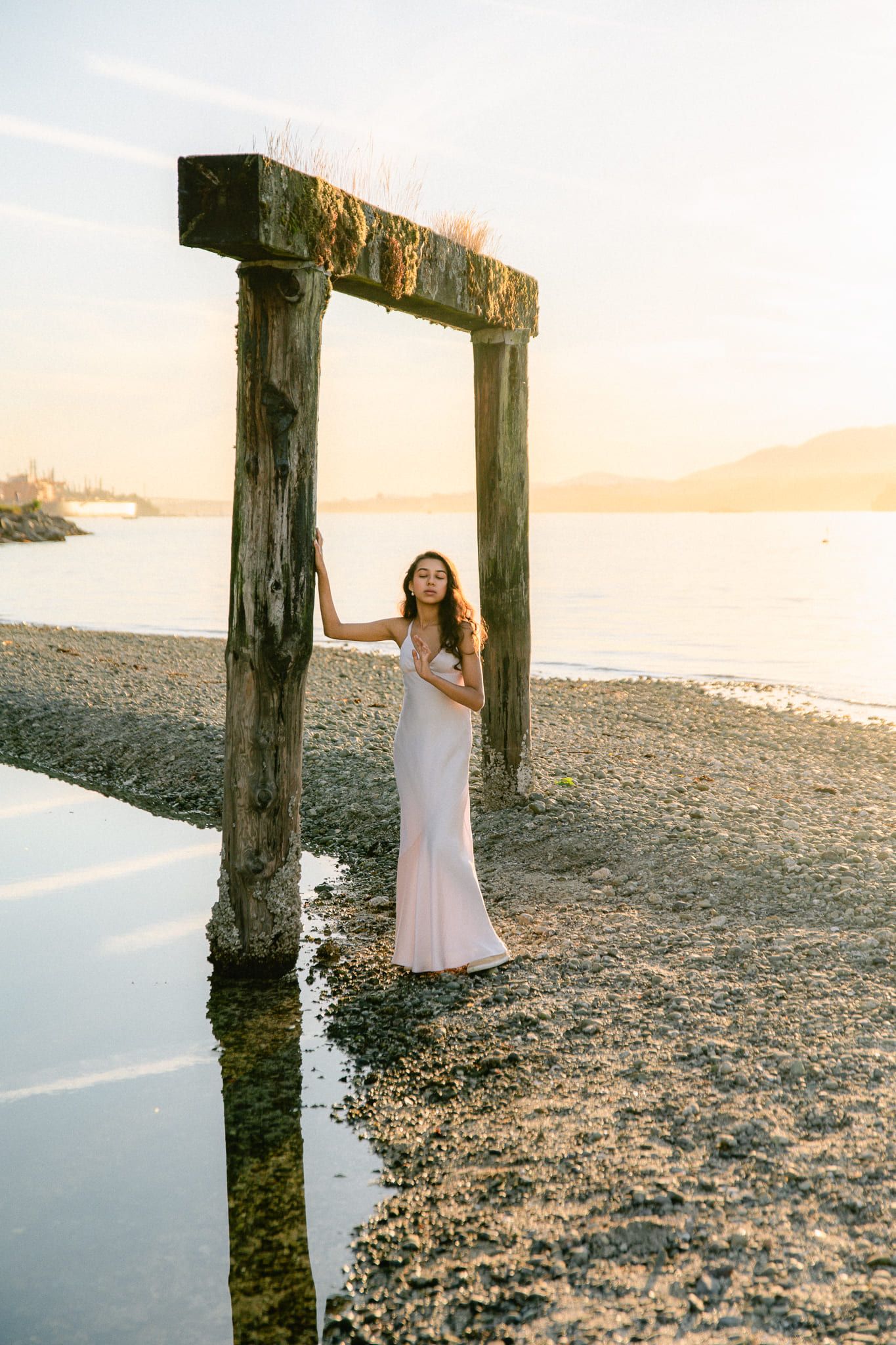 Creative portrait photography on a beach at sunset, young woman in a pale gown standing by old pier posts