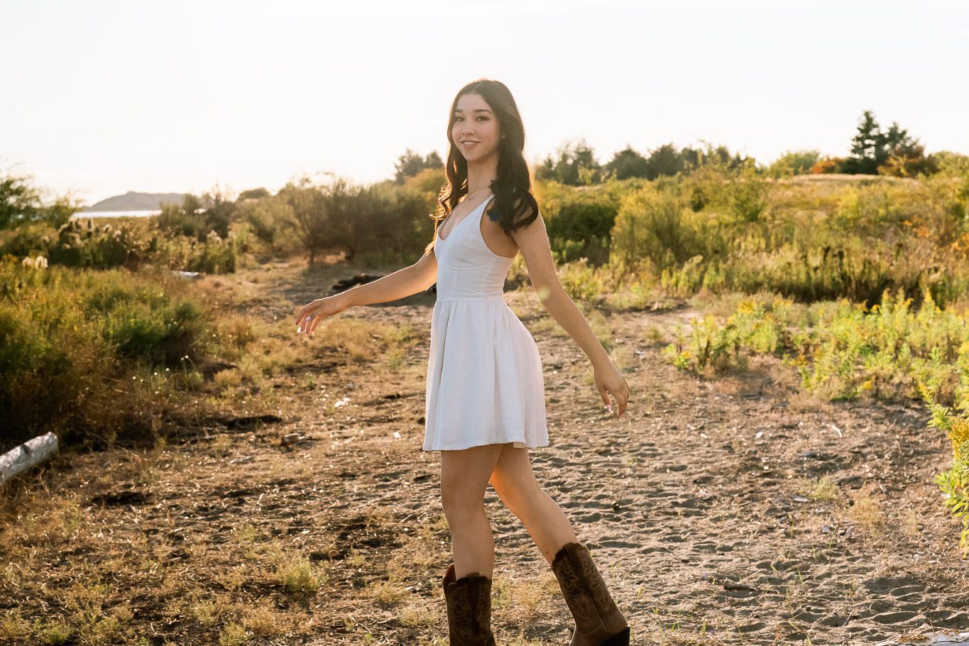 Beach portrait photo at sunset, young woman in a white dress and cowboy boots walking on a beach