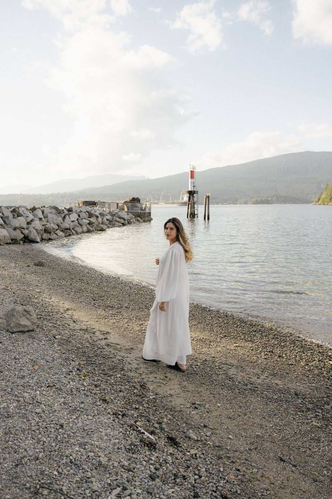 Intimate portrait photography, a woman in a flowing white robe looks back over her shoulder on a curving pebble beach