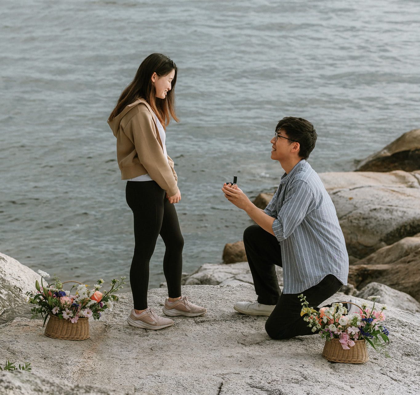 Vancouver engagement photographer, a man kneels on the rocks at the water's edge holding out a ring box to his partner, two baskets of flowers beside them