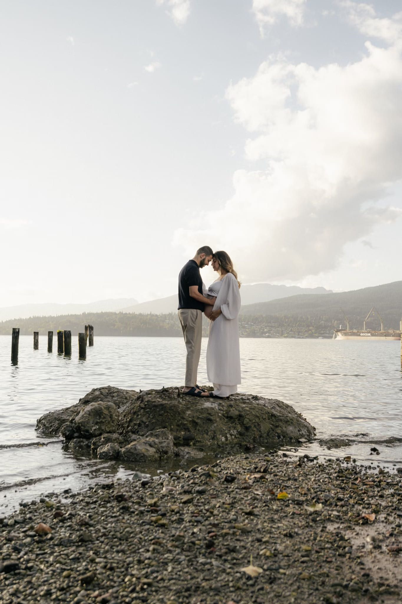 Couple embracing on a rock at the water's edge with old pilings and mountains behind