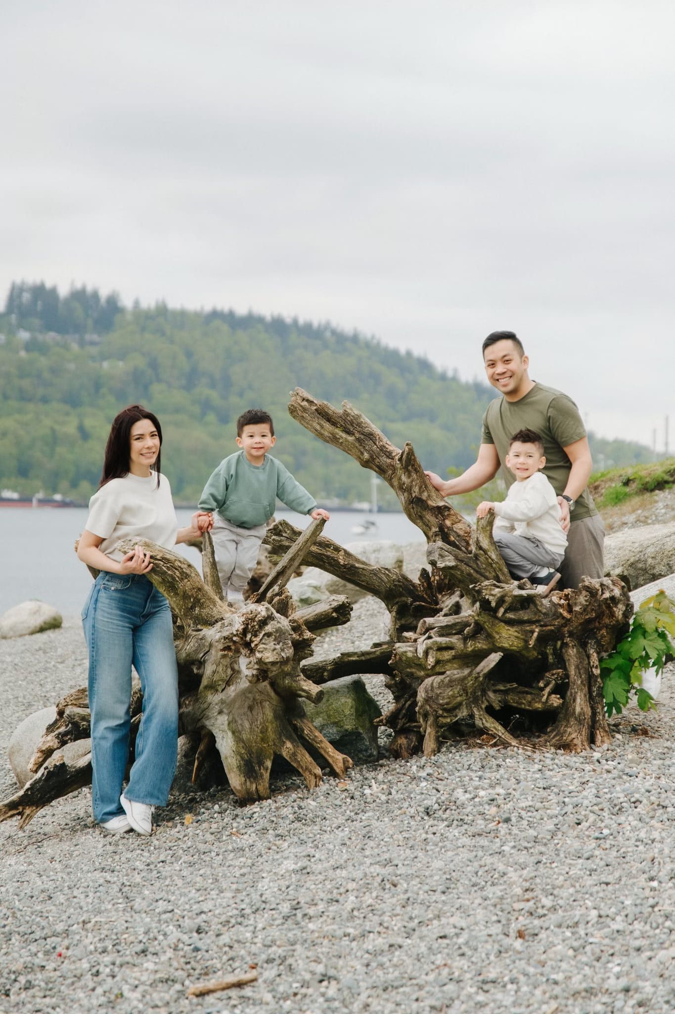 Family of four posing on driftwood on a pebble beach with a forested hill behind