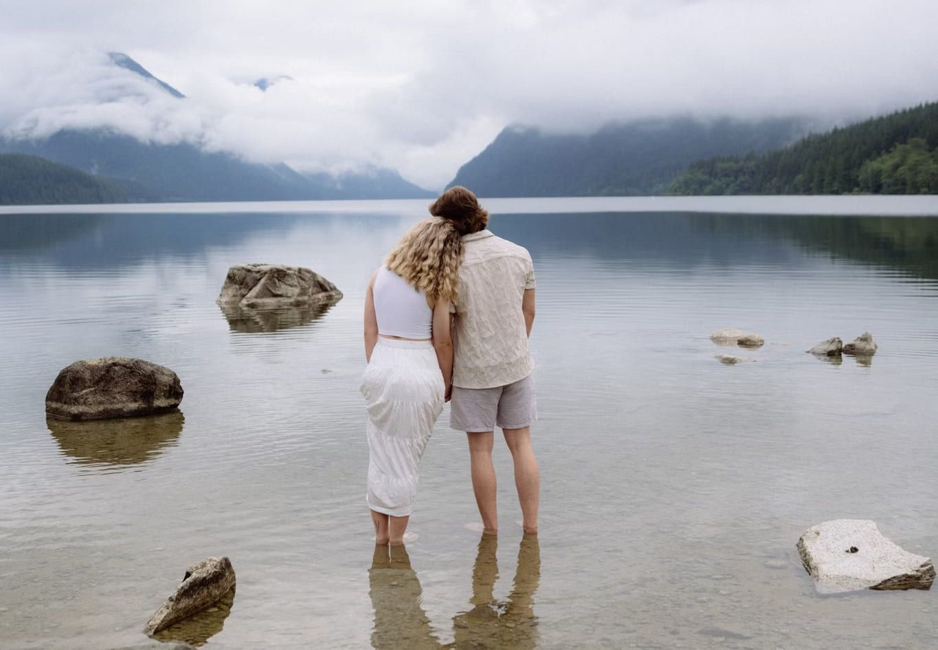 Couple standing ankle-deep in a still mountain lake beneath low cloud