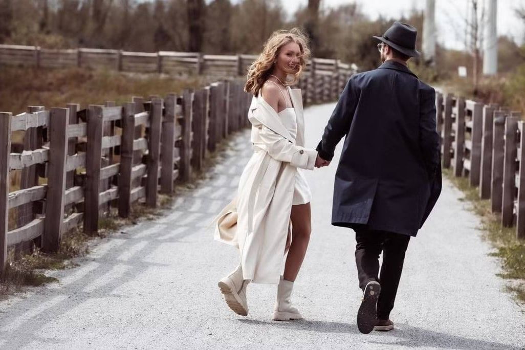 Couples photoshoot in Langley, couple walking hand in hand down a country lane beside a wooden fence, she in a white dress, he in a dark coat and hat