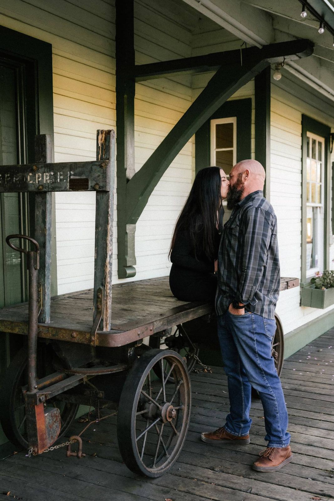 Creative engagement photos, a couple kisses beside an antique wooden cart on the porch of a white clapboard building