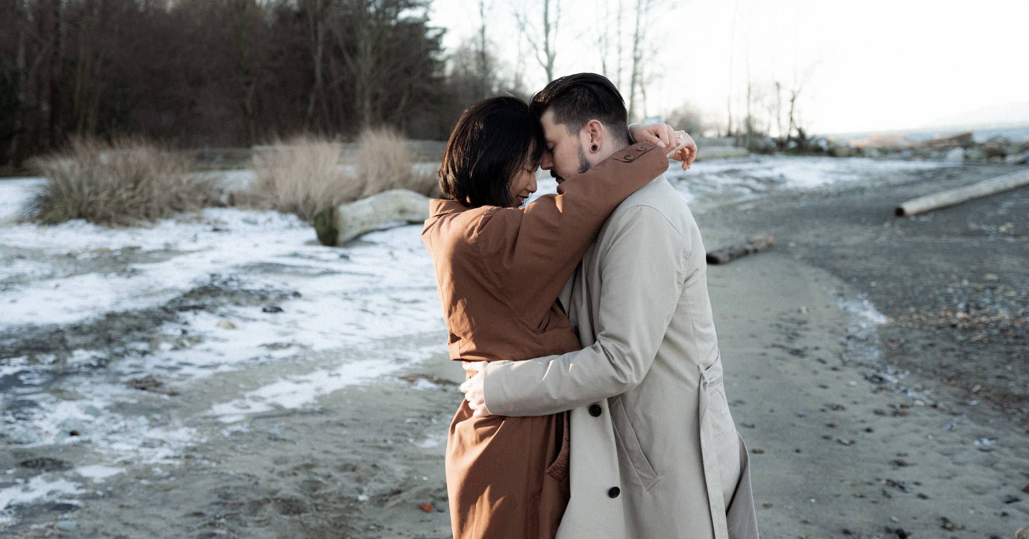 Couple in trench coats embracing forehead to forehead on a frosty winter beach