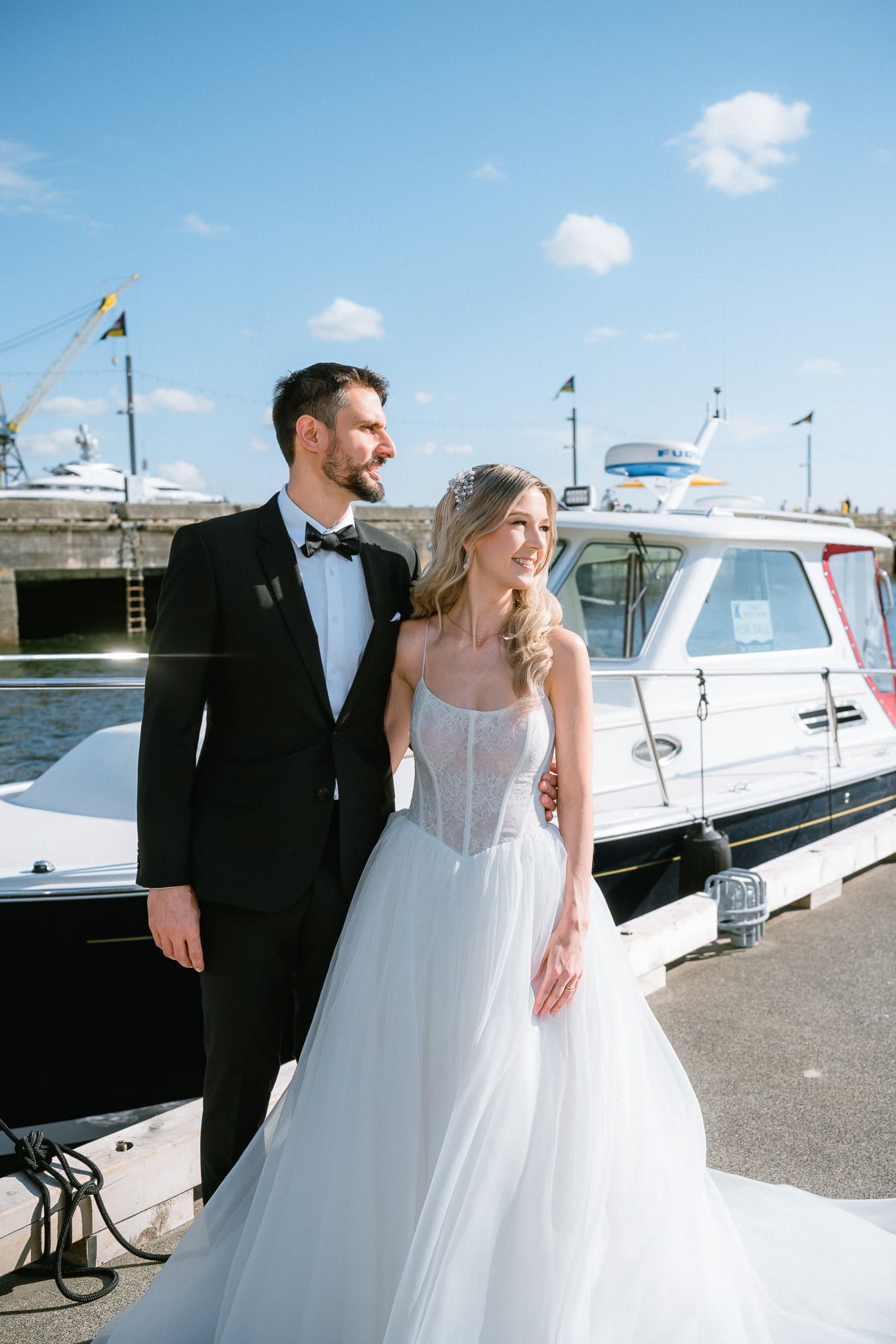 Professional wedding pictures, bride in a tulle gown and groom in a tuxedo stand beside a boat at the marina, blue sky and masts behind