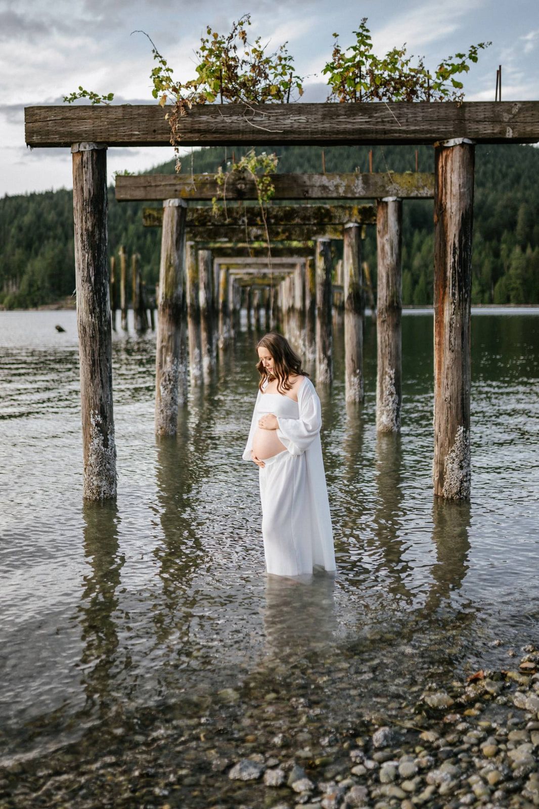 Outdoor maternity photoshoot, an expectant mother in a white gown stands in shallow water beneath rows of old pier beams