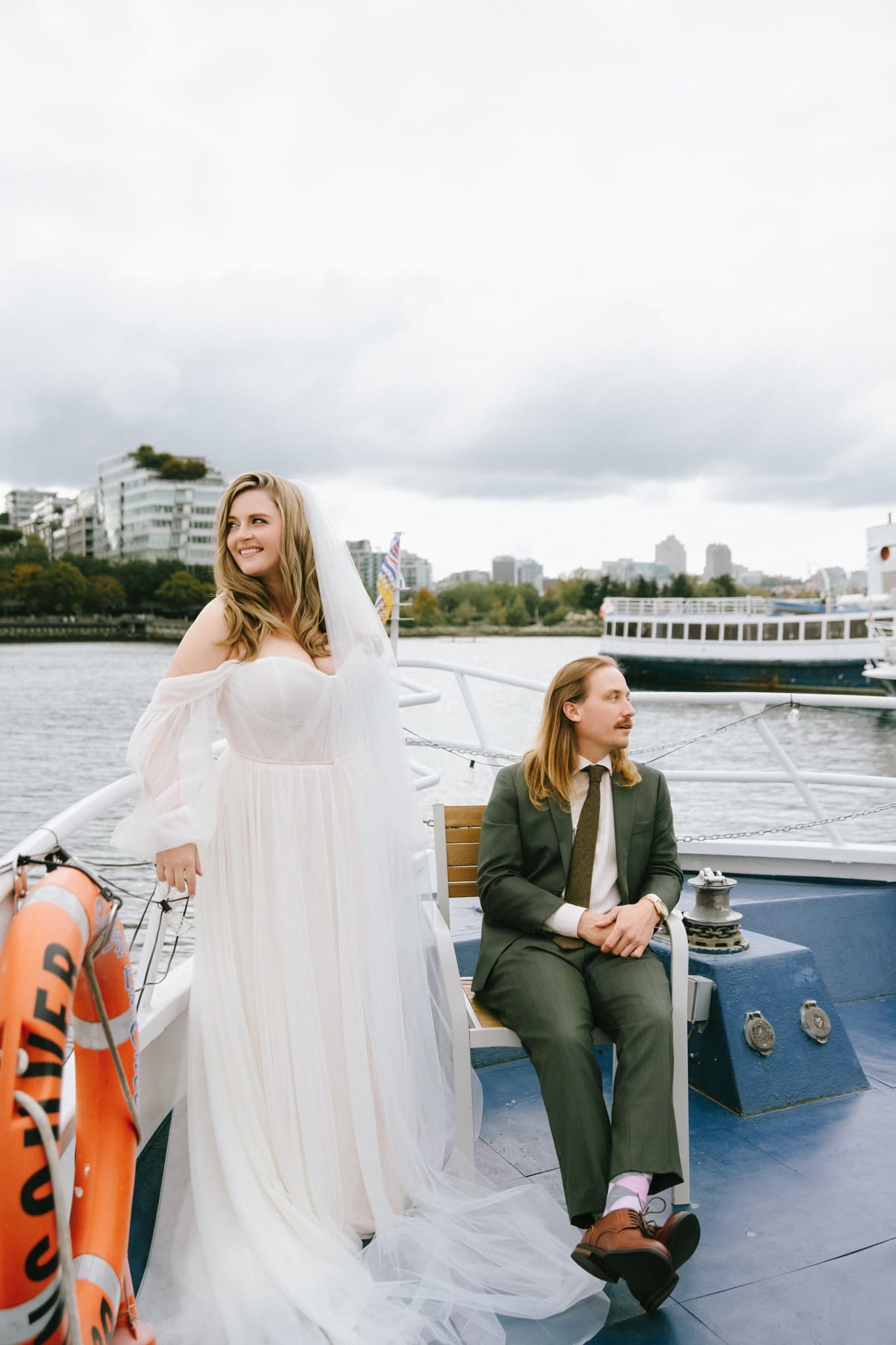 Wedding photos, bride in a tulle gown and veil and groom in a green suit on the deck of a boat in the harbour, the city skyline behind them