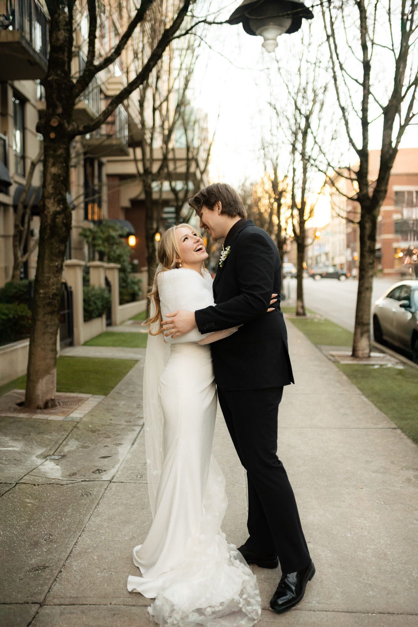 Intimate wedding photography, bride and groom embracing on a city sidewalk lined with trees