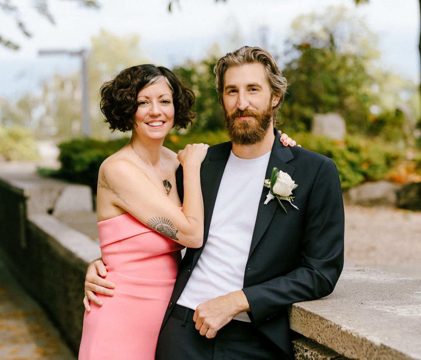 Vancouver elopement photographer, newlyweds lean on a stone ledge under the trees, she in a coral off-the-shoulder dress, he in a black suit, both smiling