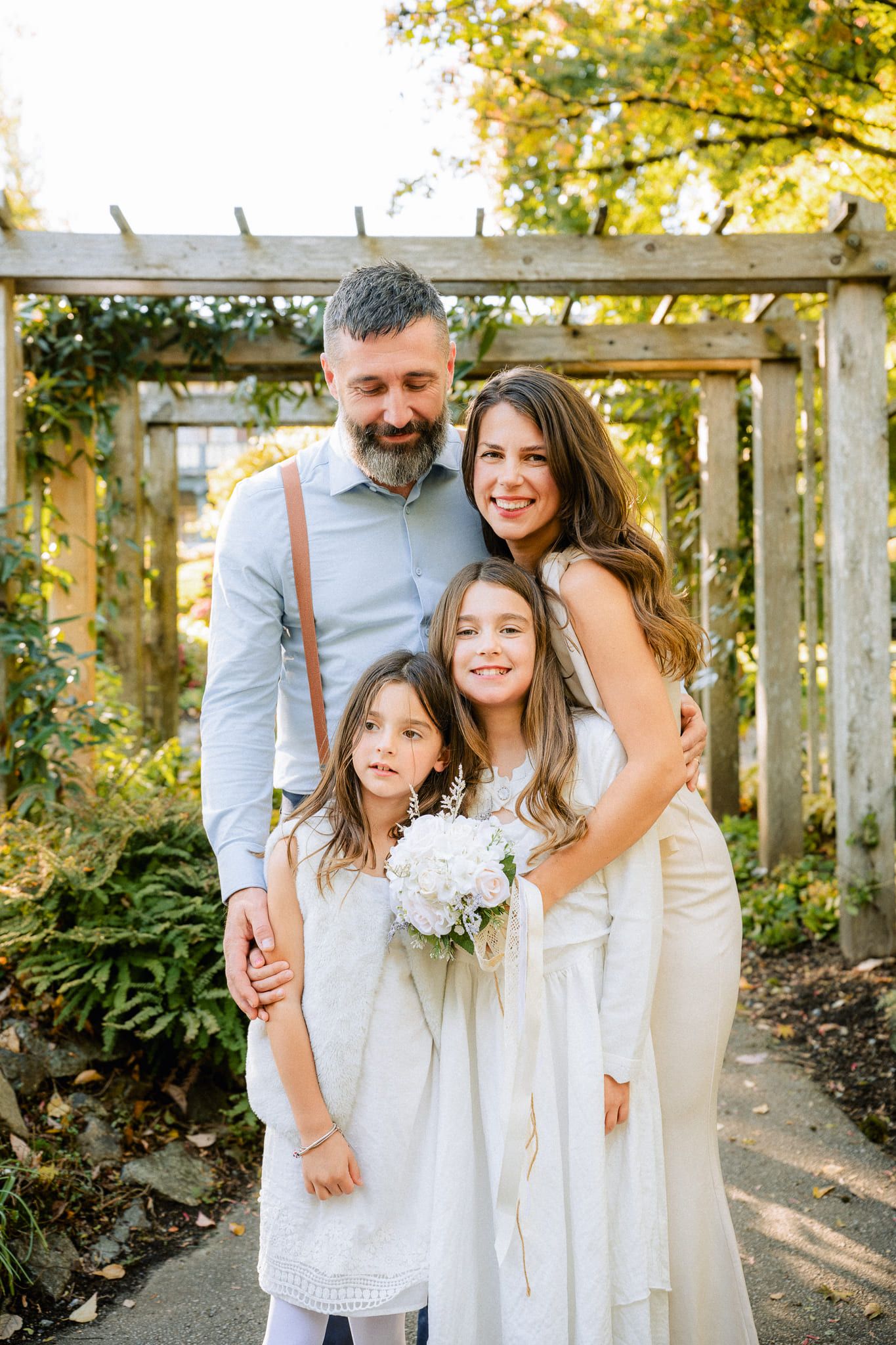 Family wedding pictures, a family of four embrace under a wooden pergola, the mother in a white gown holding a bouquet, two daughters in white in front