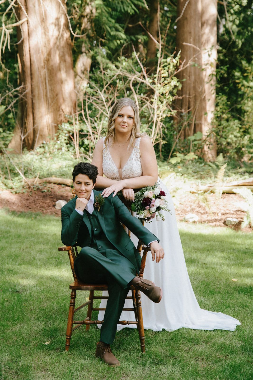 Intimate wedding photos among tall trees, newlywed in a green suit seated on a wooden chair, the bride standing behind