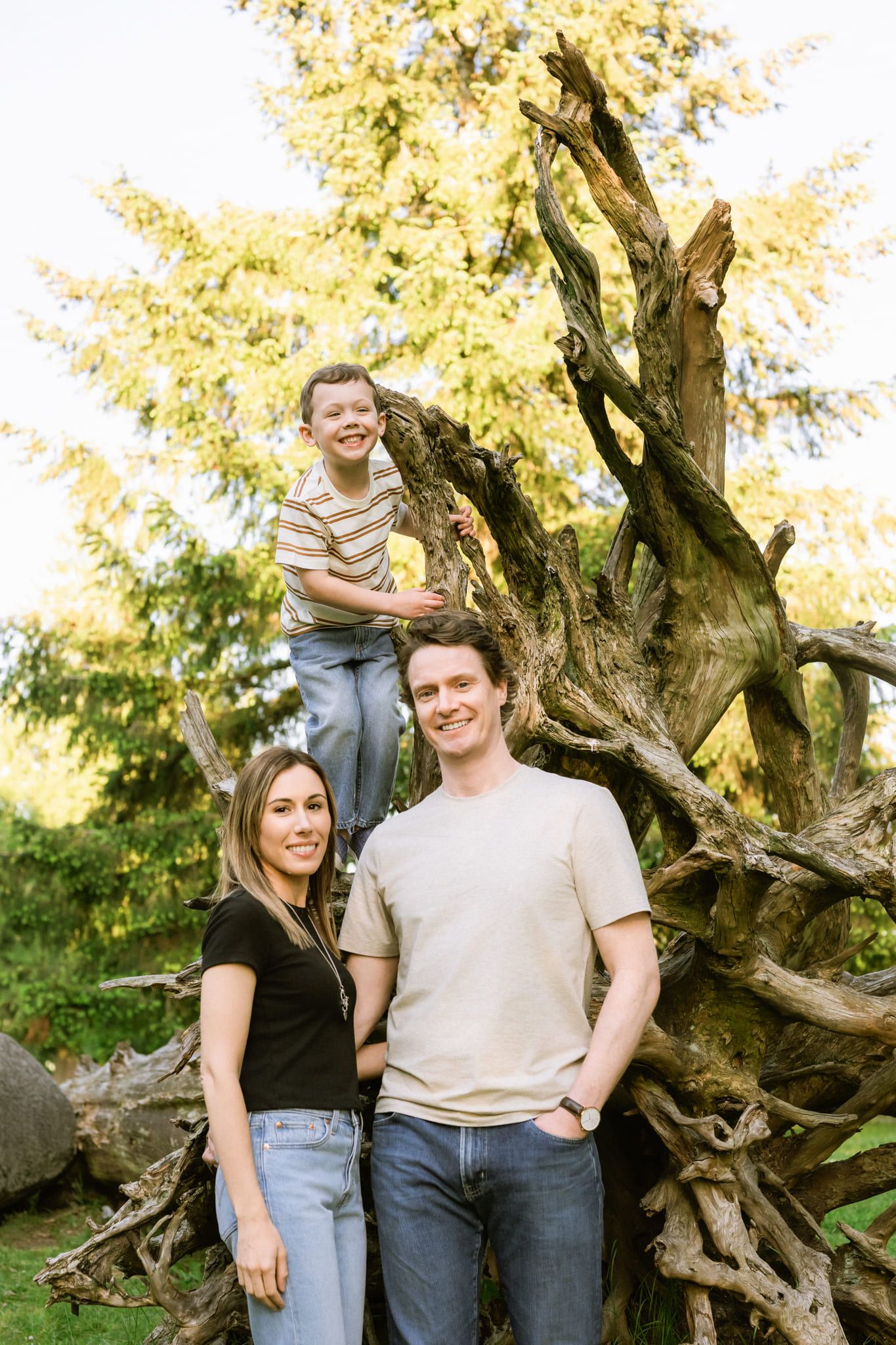 Small family photo, parents standing by a driftwood stump while their son climbs it