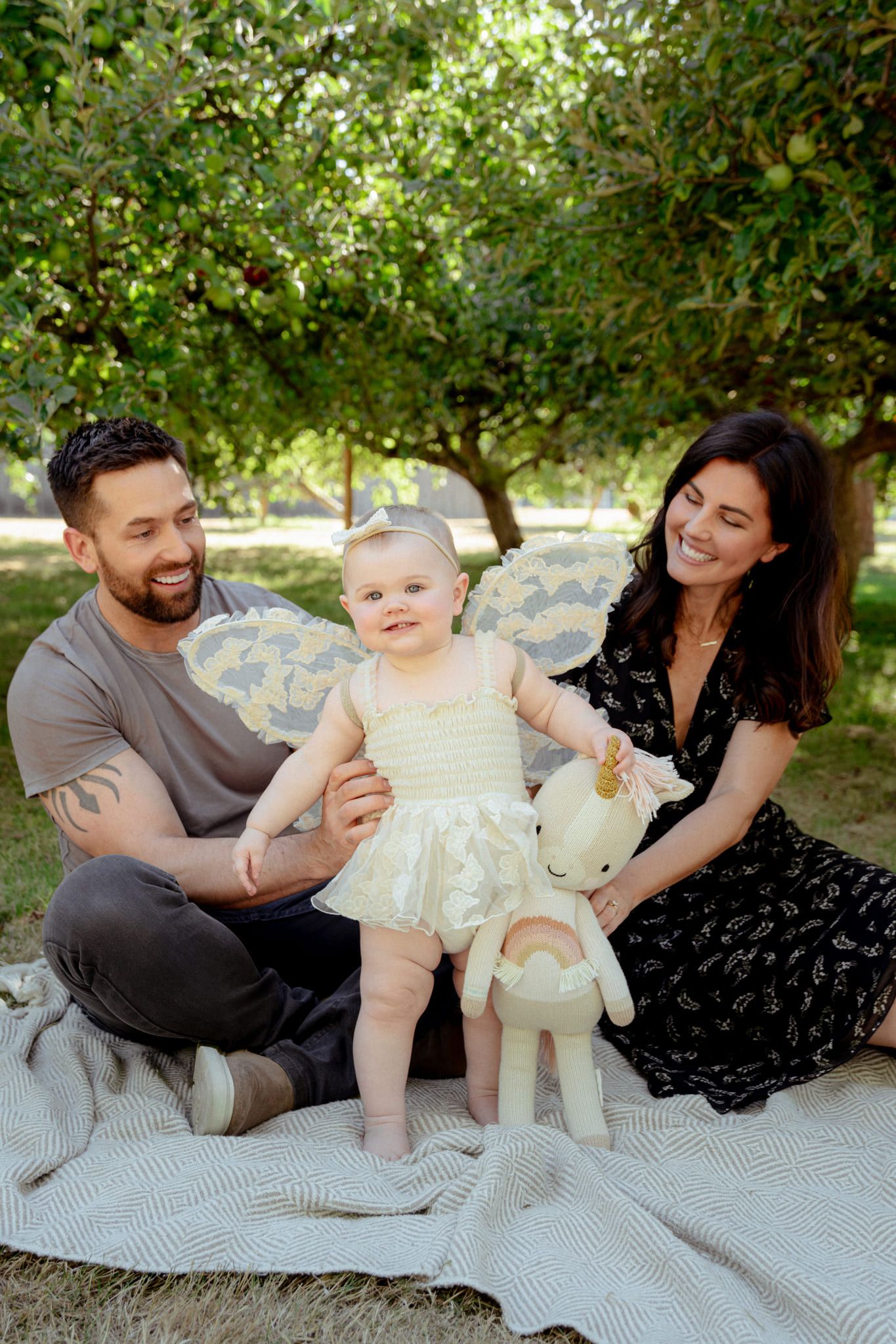 Modern family photography, parents sitting on a blanket under a tree as their baby girl in a tulle dress stands between them
