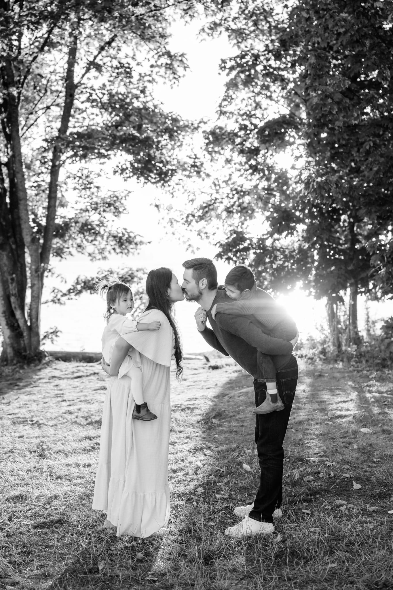 Black and white family picture among the trees, parents kissing while carrying their kids on their backs