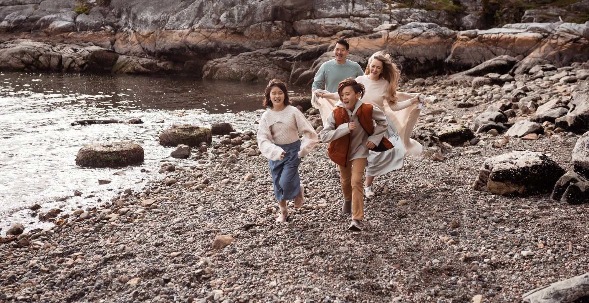 Family of four running along a rocky beach, the children laughing ahead of their parents