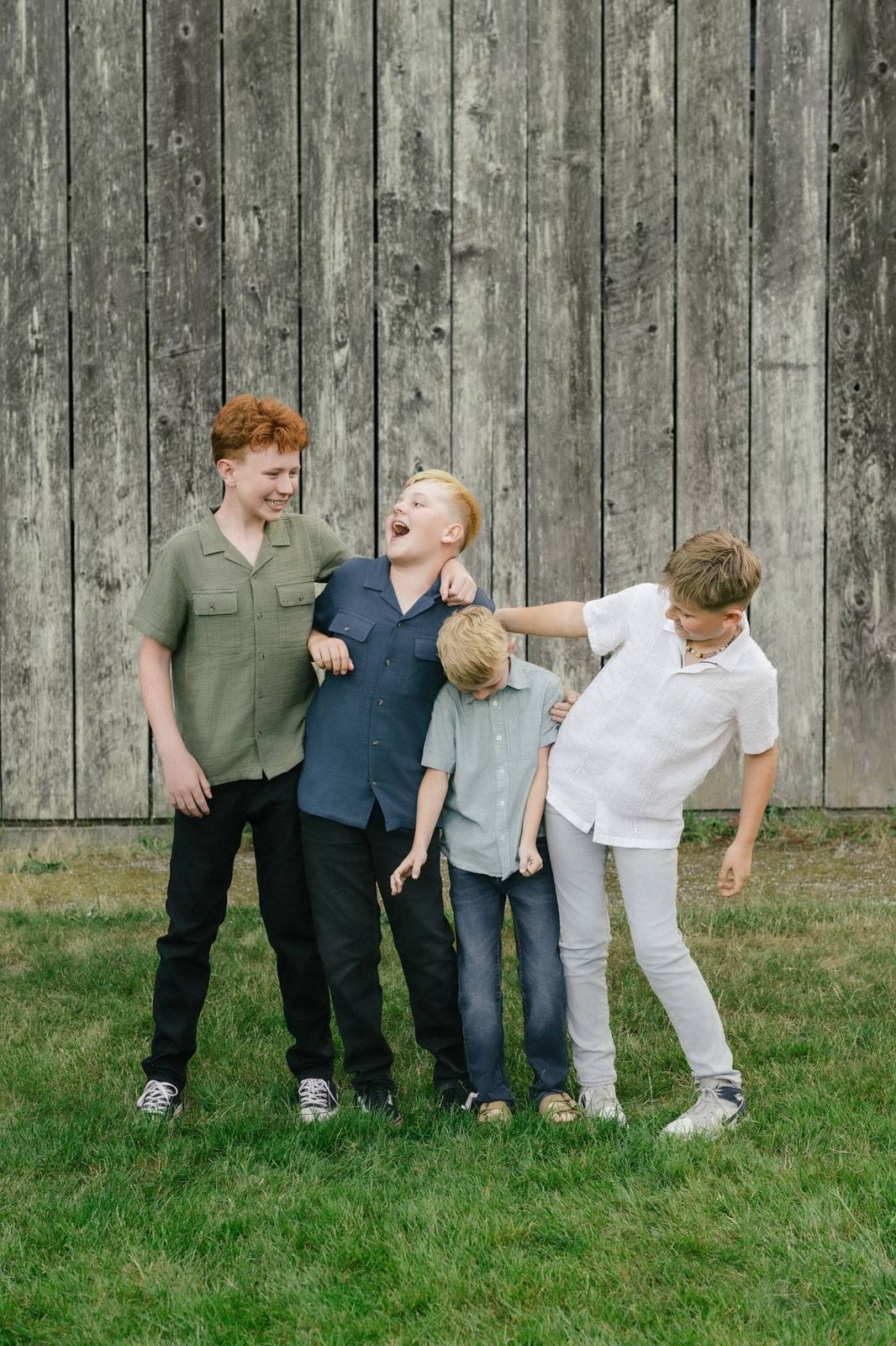 Four boys laugh and jostle each other in front of a weathered wooden barn wall