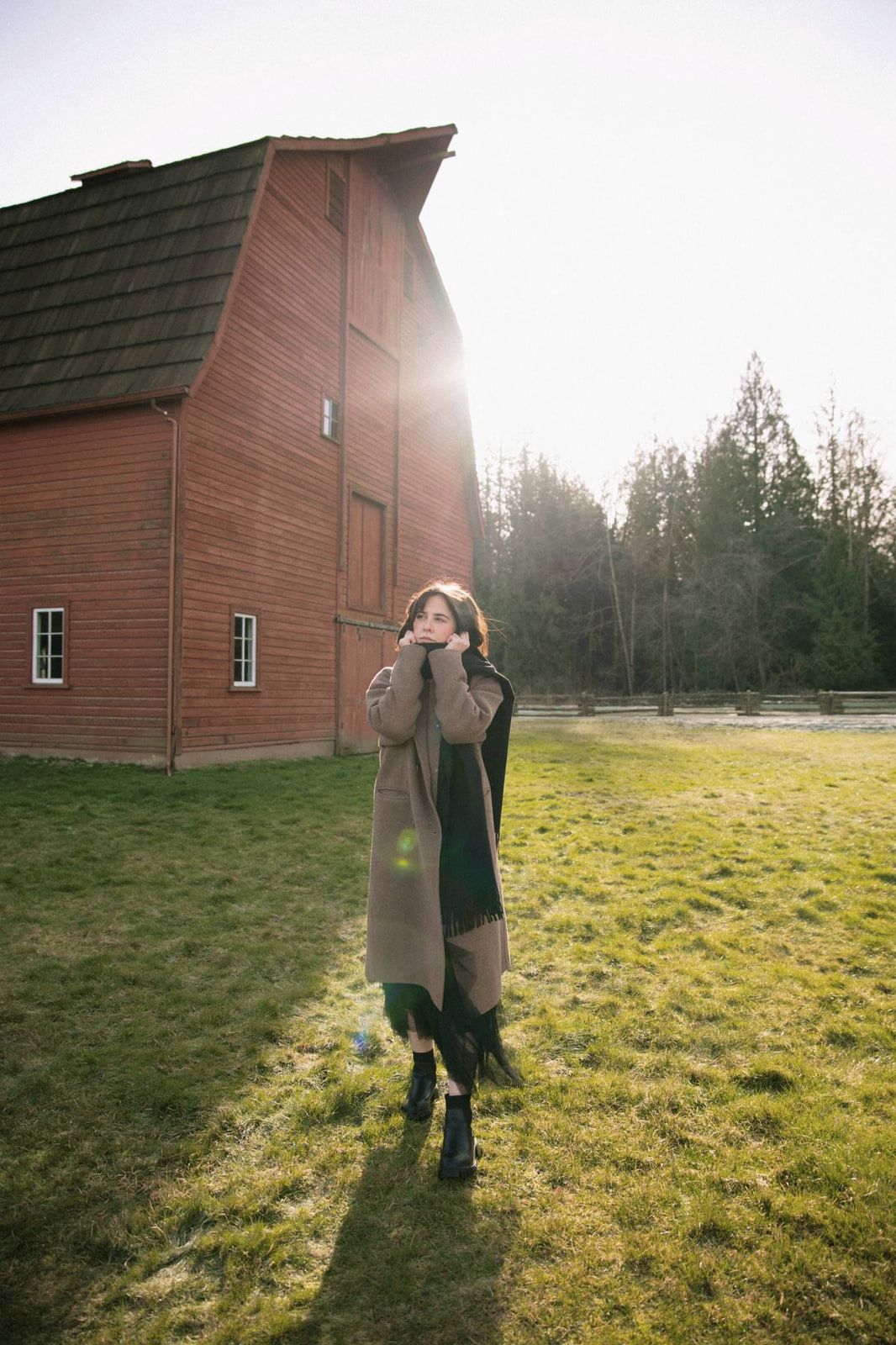 Professional portrait photo in bright sunlight, a woman in a long coat stands on the grass beside a red barn