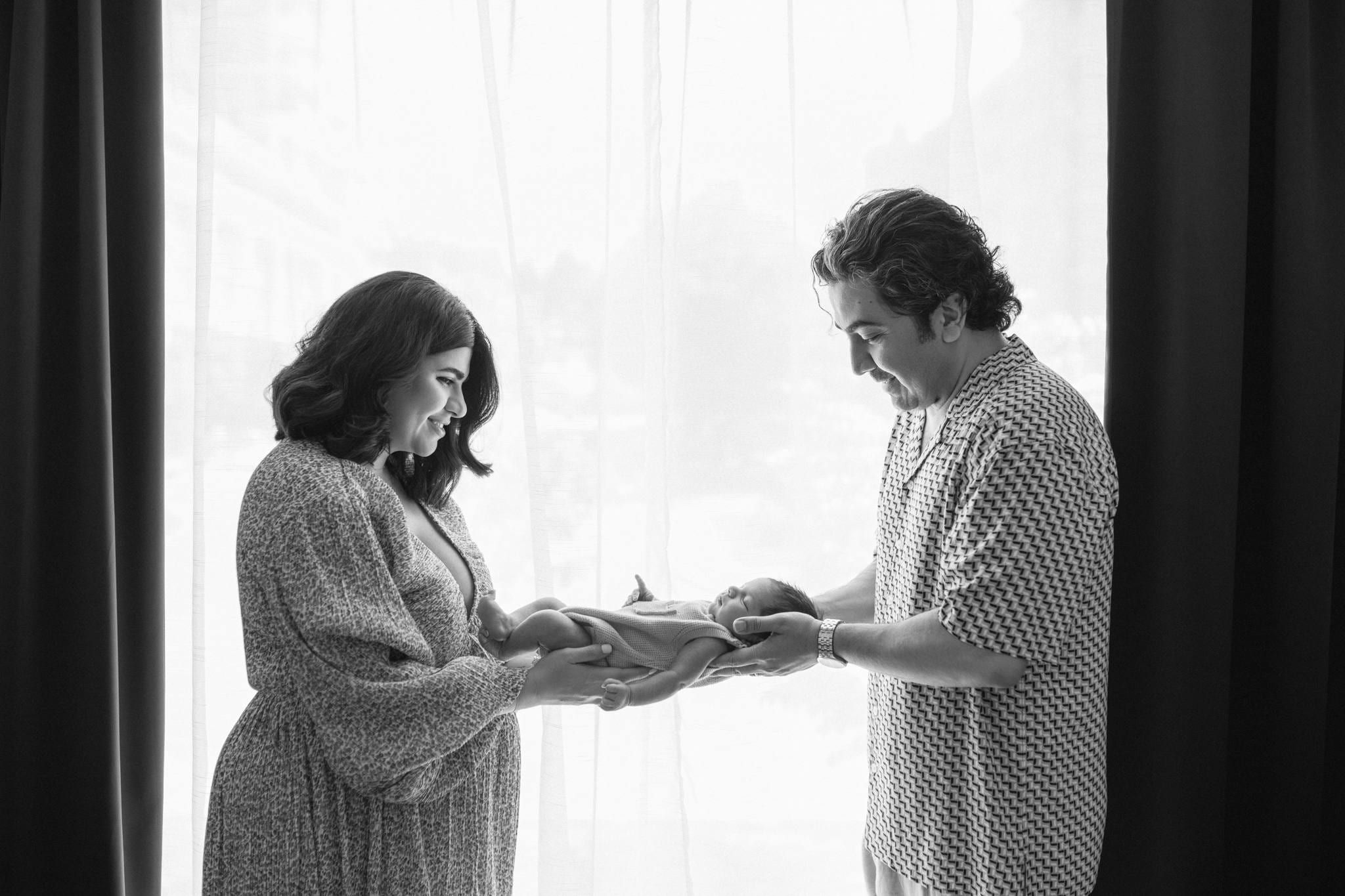 Black-and-white photo of parents holding their newborn between them in front of a sheer window
