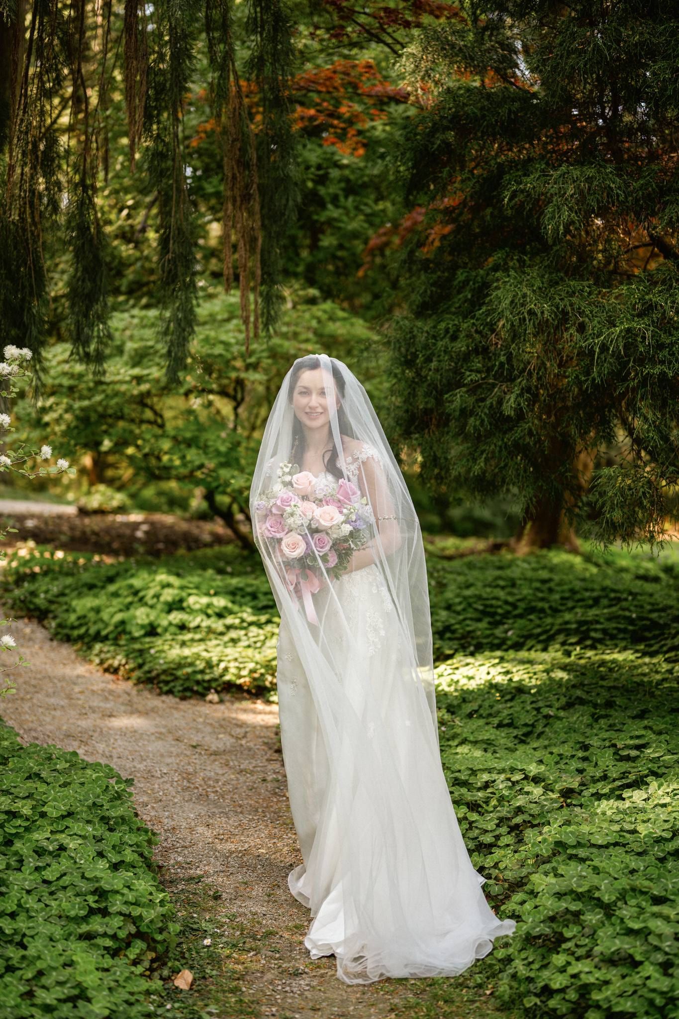 Bride beneath her veil holding a pink bouquet on the garden path