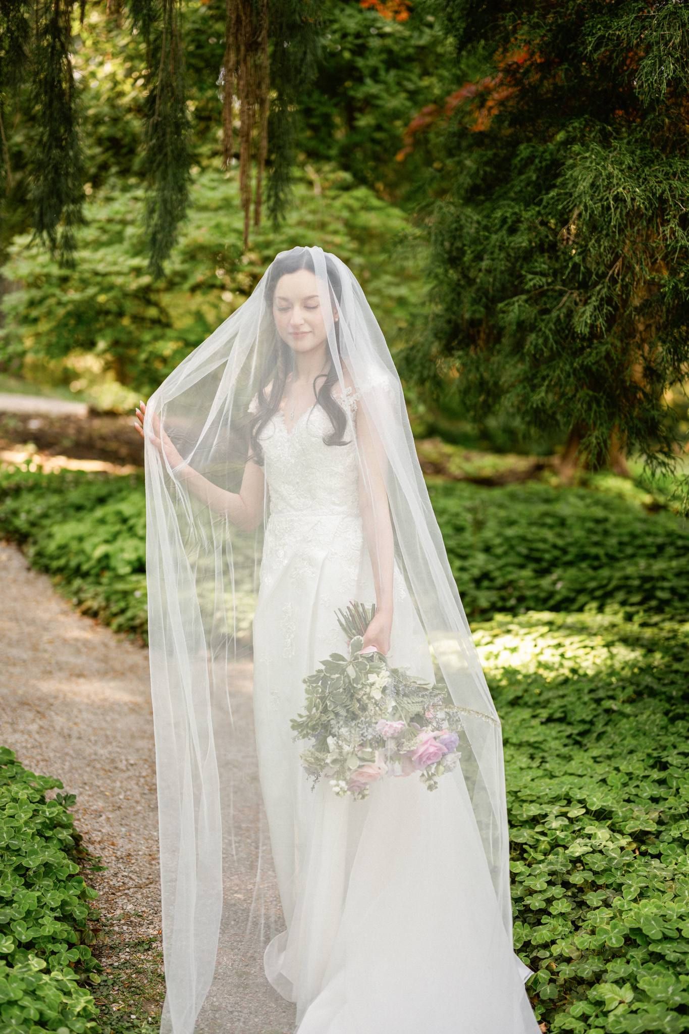 Bride under a long veil holding her bouquet by the garden path