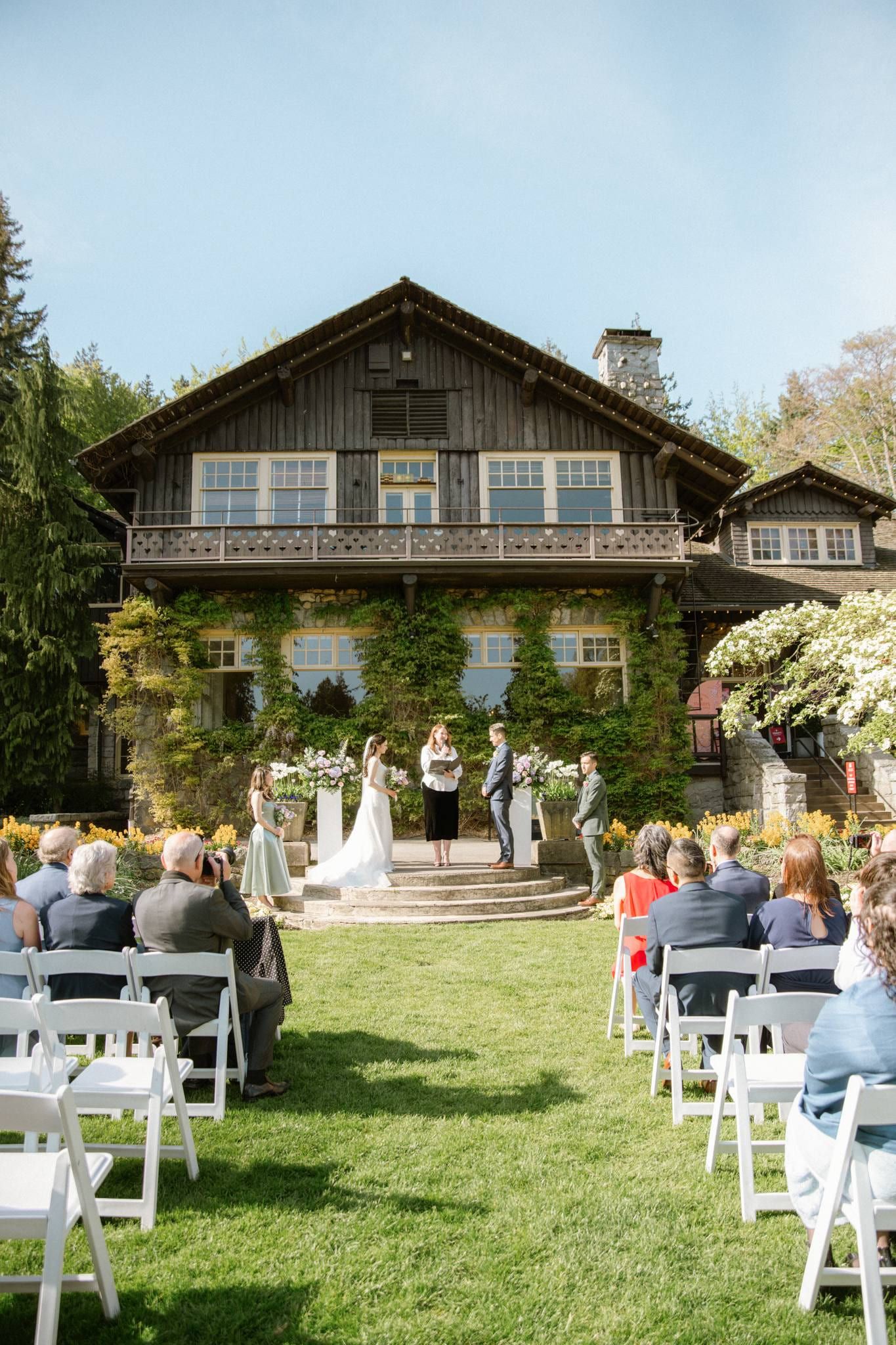 Angela and Jason exchanging vows before the stone lodge as guests look on from white chairs