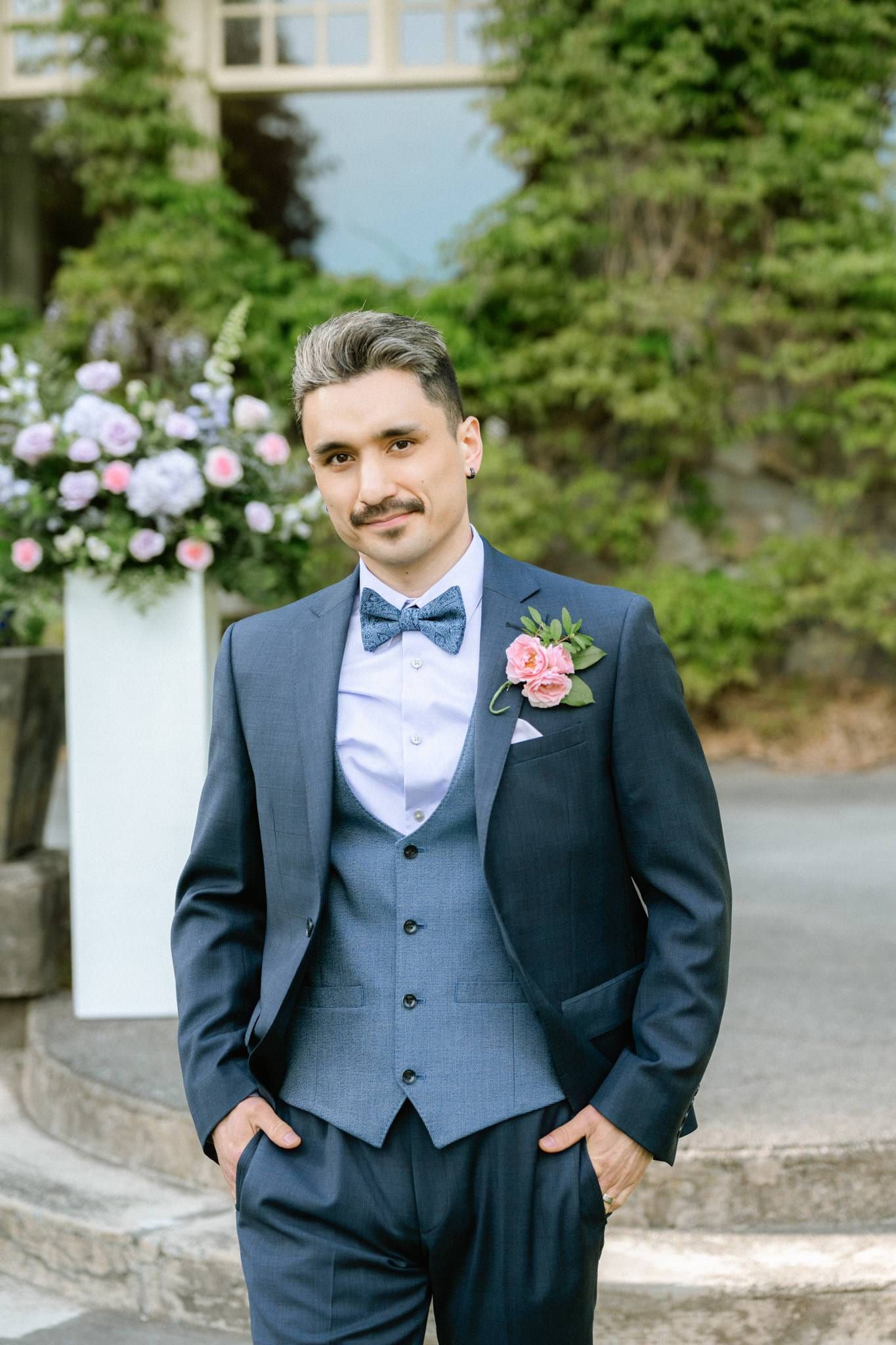 Groom in a blue suit and bow tie beside the floral arch